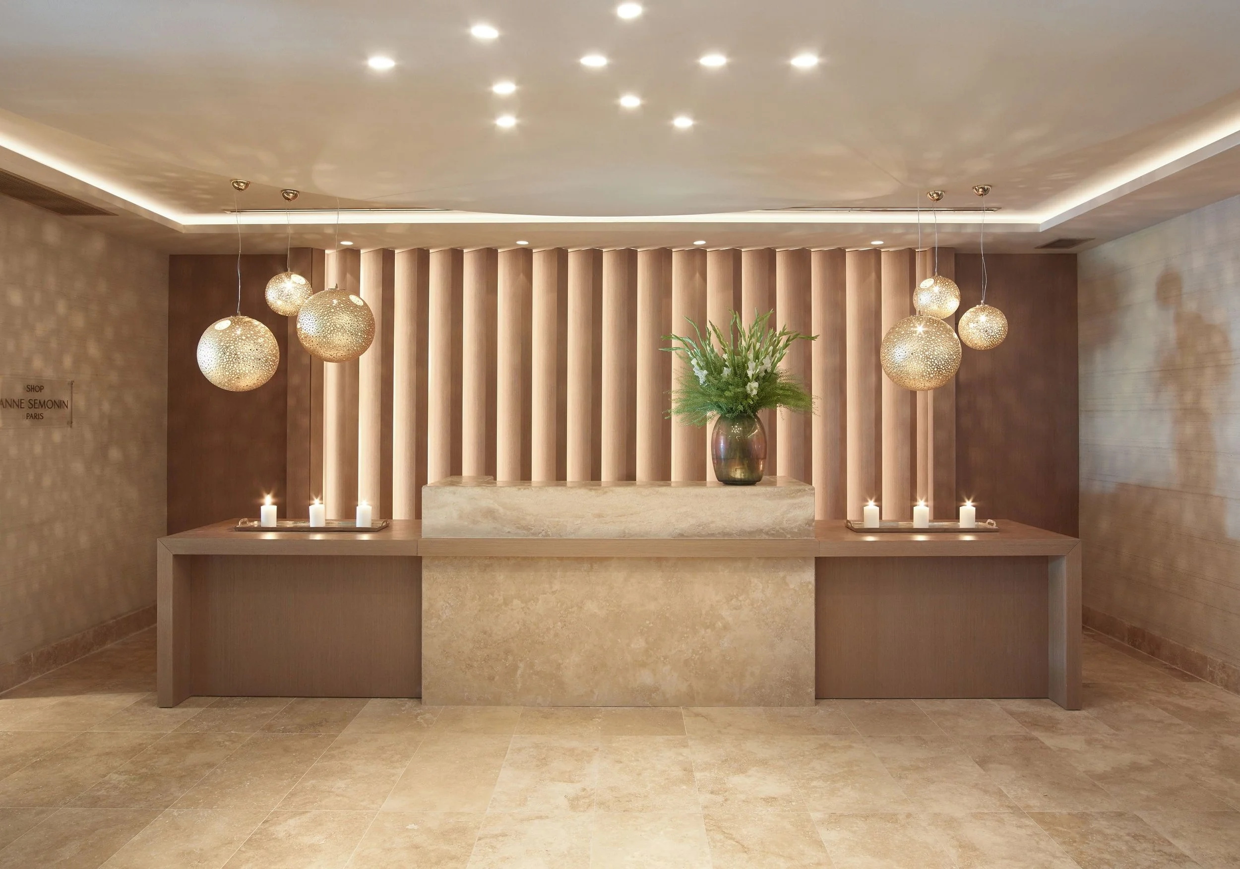 Elegant lobby with a beige stone reception desk, a large vase with green plants on top, illuminated hanging pendant lights, and a backdrop of wooden vertical slats, with candles on the sides.