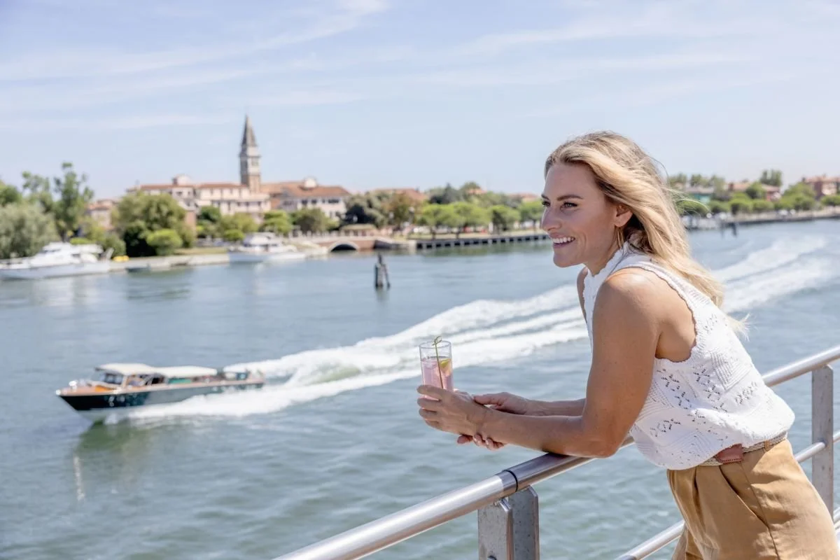 A woman leaning on a railing by a river, holding a drink, smiling, with boats and a cityscape in the background.