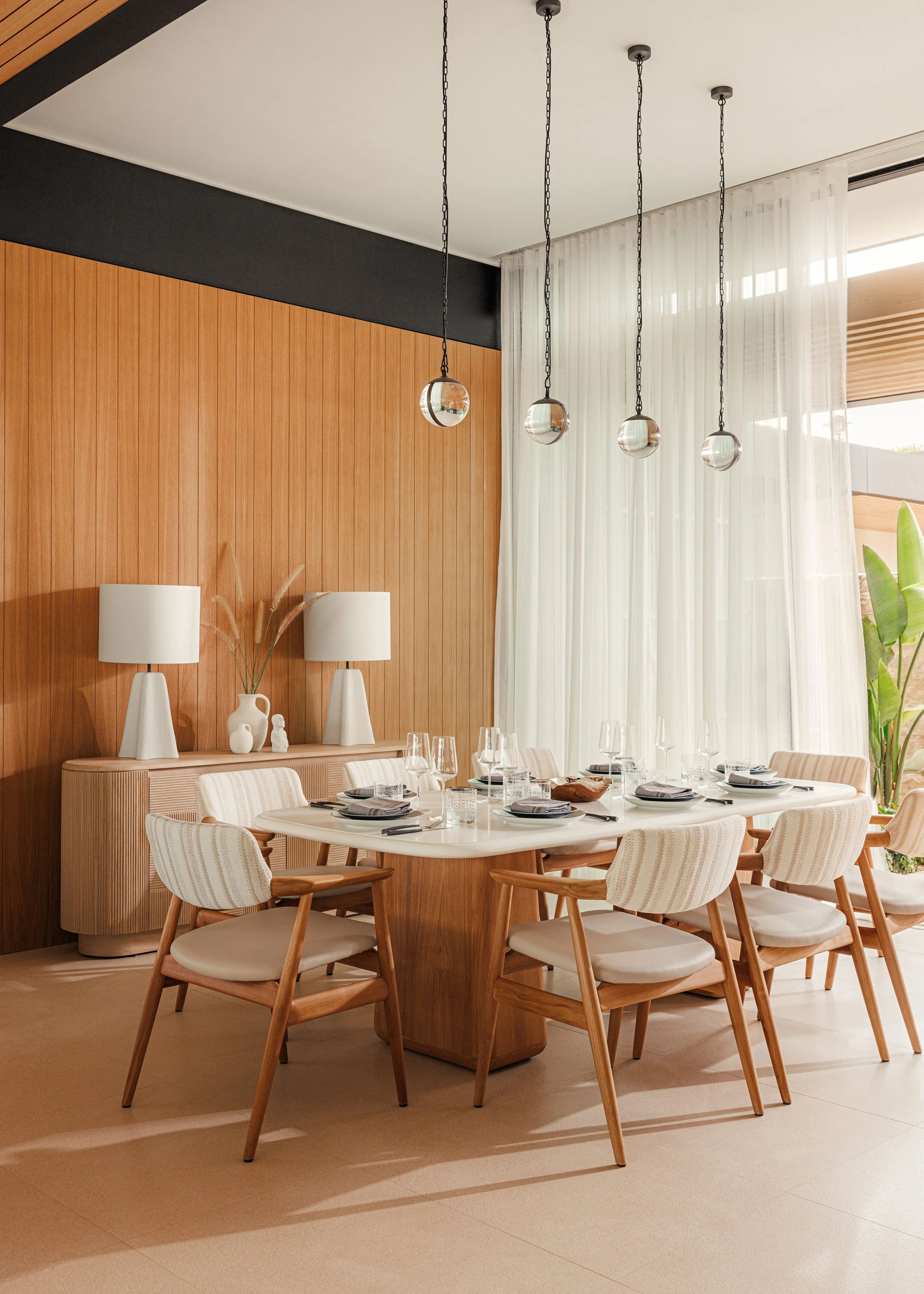 A modern dining room with a round white table set for six, surrounded by wooden and white upholstered chairs. Above the table, four round glass pendant lights hang from black chains. The room has wood-paneled walls, a sideboard with white lamps and vases, sheer white curtains, and a large window letting in natural light.