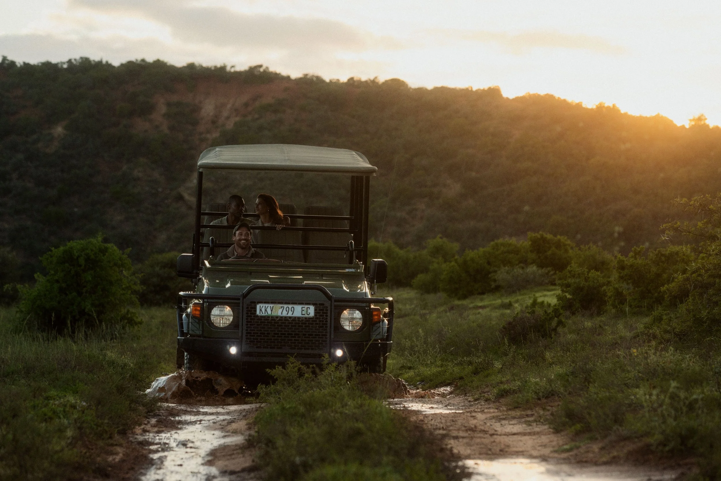 A safari vehicle with three people in it driving through a muddy, grassy landscape during sunset with hills in the background.
