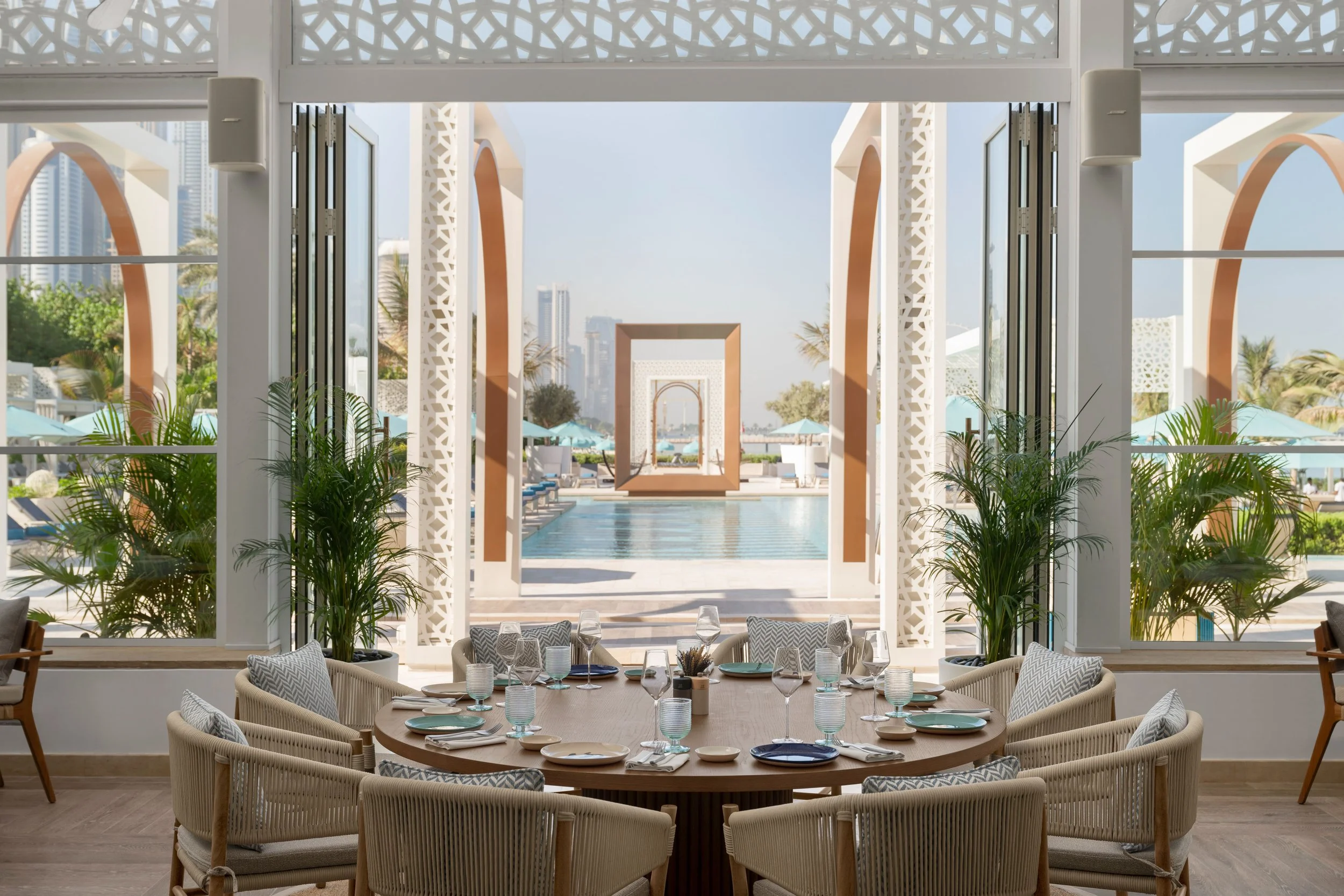 A round dining table set with white plates, wine glasses, and water glasses, surrounded by woven chairs with patterned cushions, inside a bright room with large windows and views of a pool and city skyline in the background.