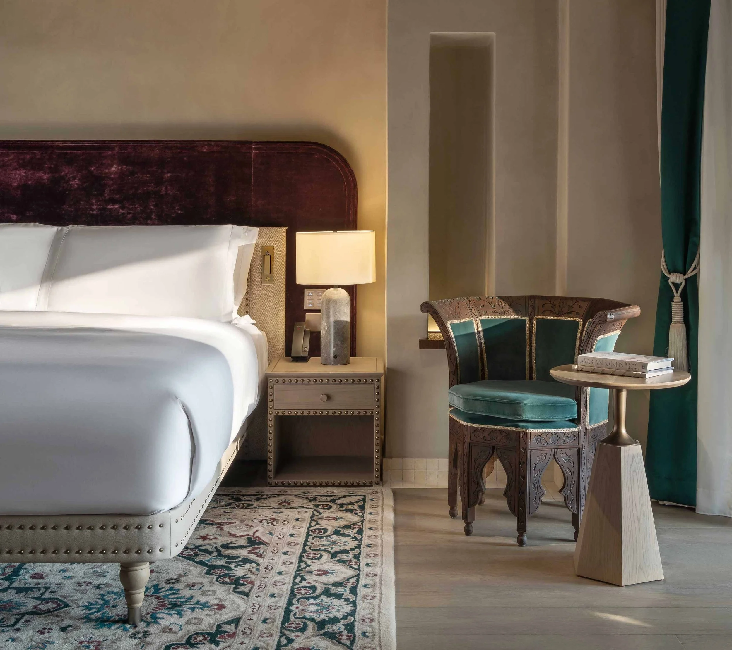 A bedroom with a white bed, a nightstand, a lamp, an armchair, a rug, and a window with curtain.