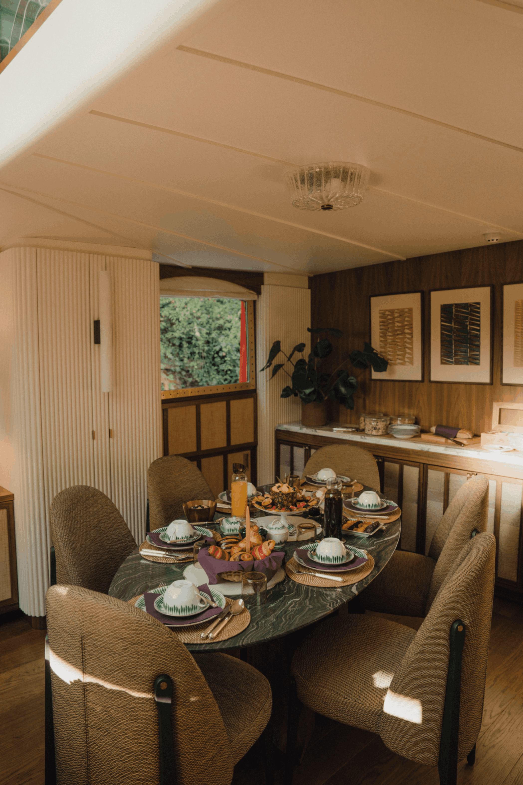 Dining room with a large round table set with plates, bowls, glasses, and a centerpiece of cookies and snacks, surrounded by upholstered chairs. The room has wooden walls, framed artwork, a window with greenery outside, and a ceiling light fixture.