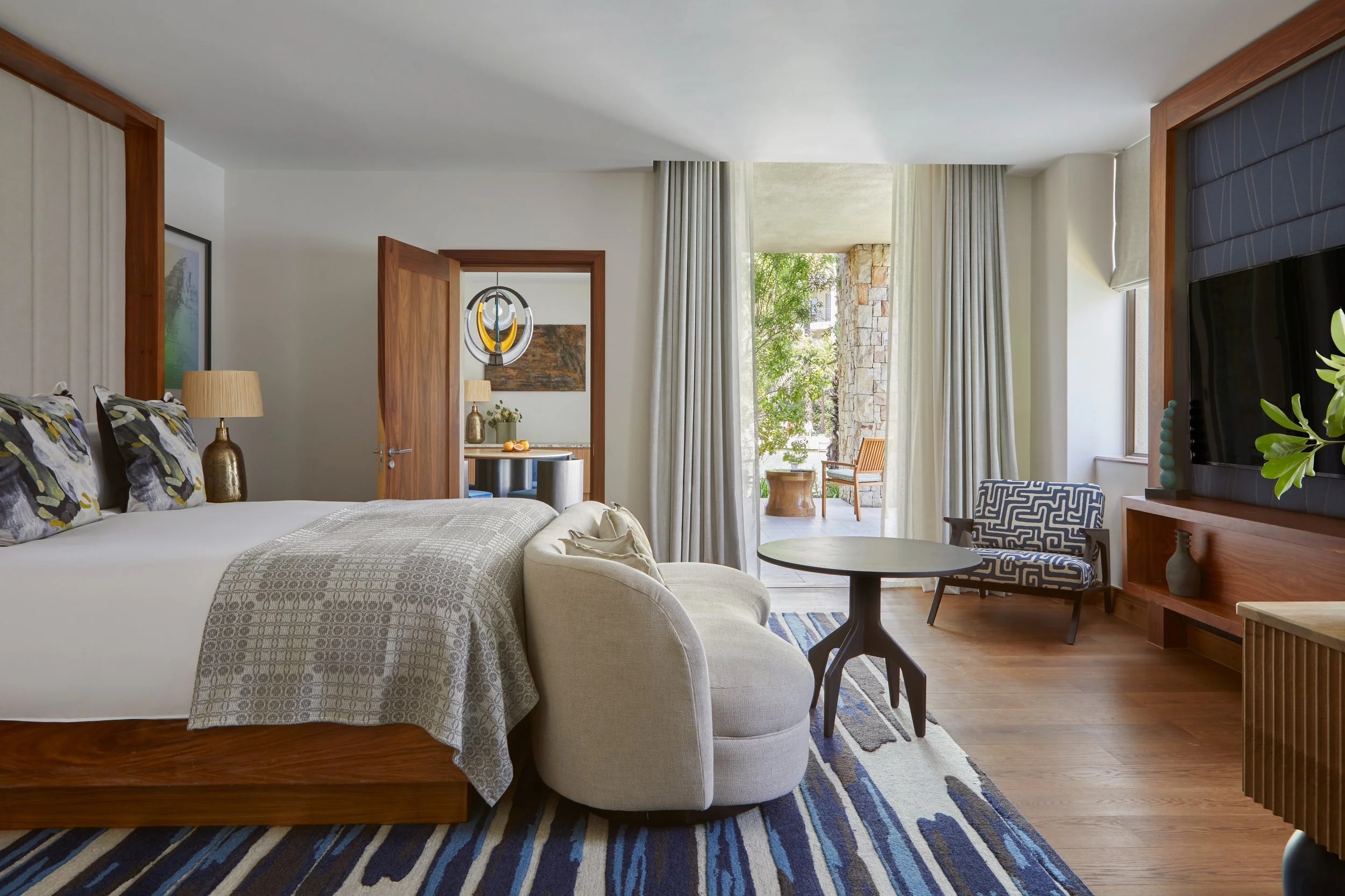 A spacious hotel room with a bed, a sitting area, and large windows showing green trees outside. The decor includes wooden accents, a striped area rug, and modern furniture like a patterned armchair and a round coffee table.