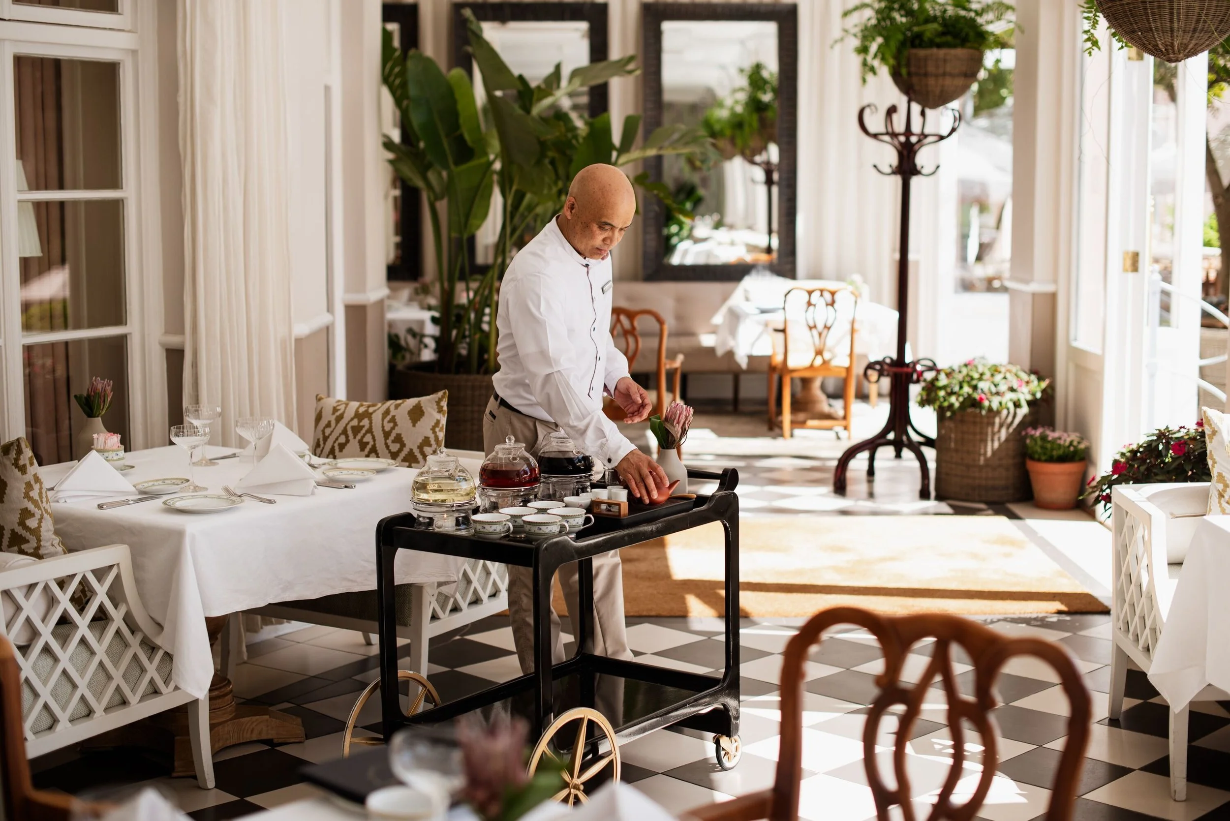 A waiter preparing drinks at a restaurant with large windows and indoor plants.