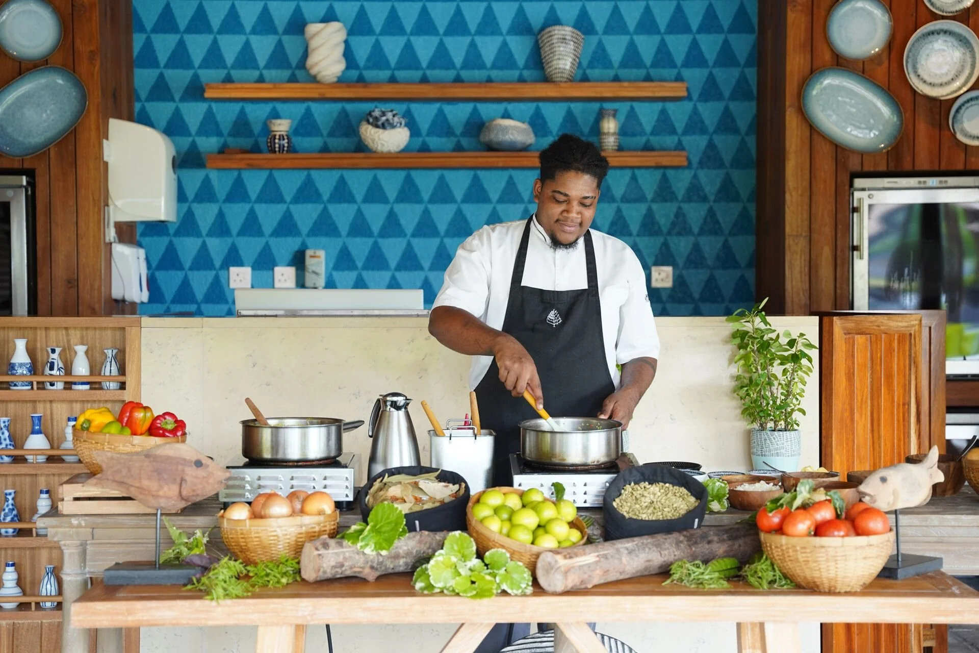 A man is cooking in a kitchen with a blue triangle-patterned wall behind him. The kitchen has wooden shelves with decorative ceramics, a potted plant, and a variety of fresh vegetables and herbs displayed on the counter.