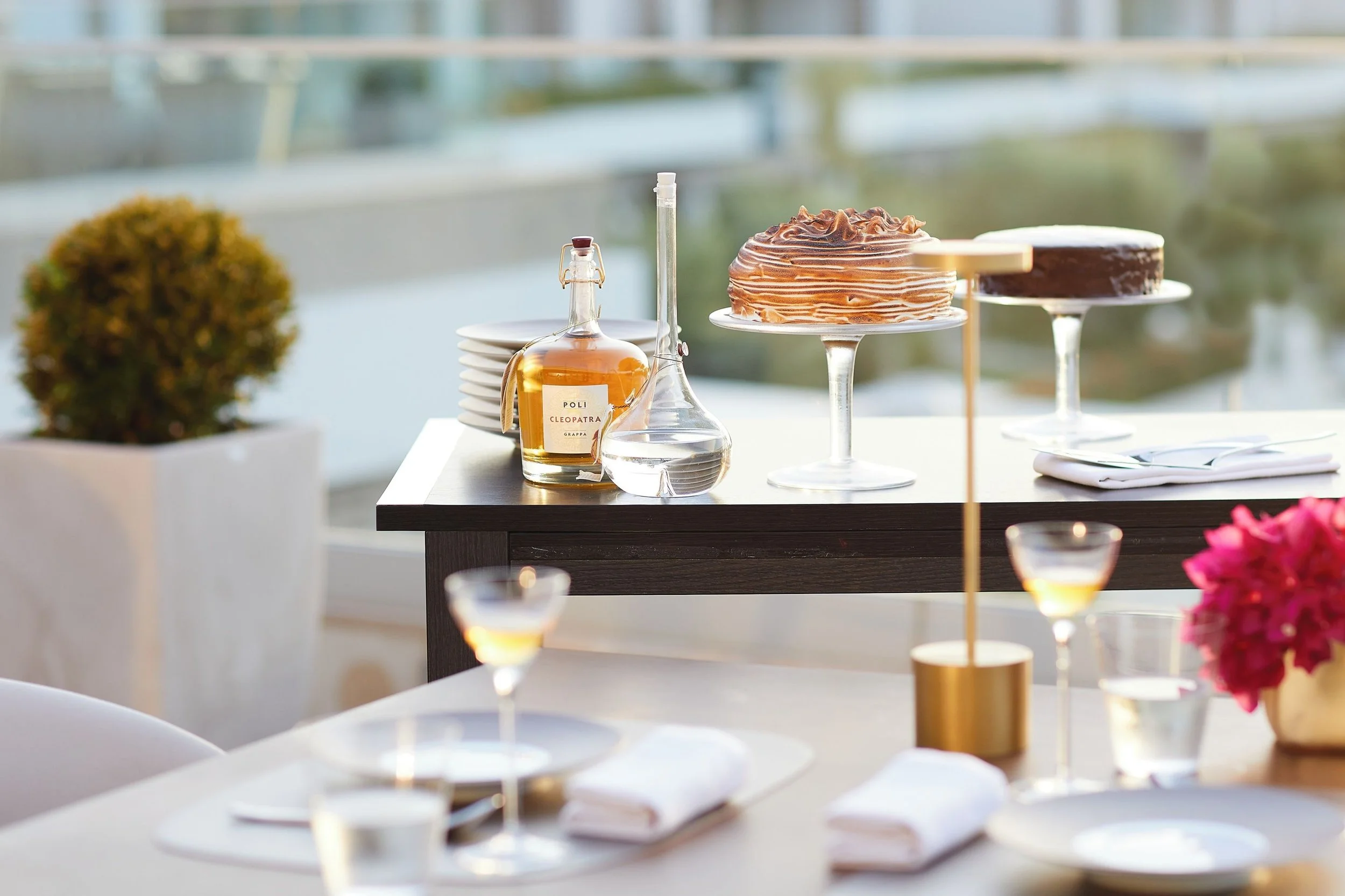A table decorated with desserts and beverages, including layered cakes on stands, a bottle of liquor labeled 'Poli Cleopatra,' white plates, gold-rimmed glasses, and a pink flower arrangement in a gold vase, with a blurred background of a balcony and greenery.