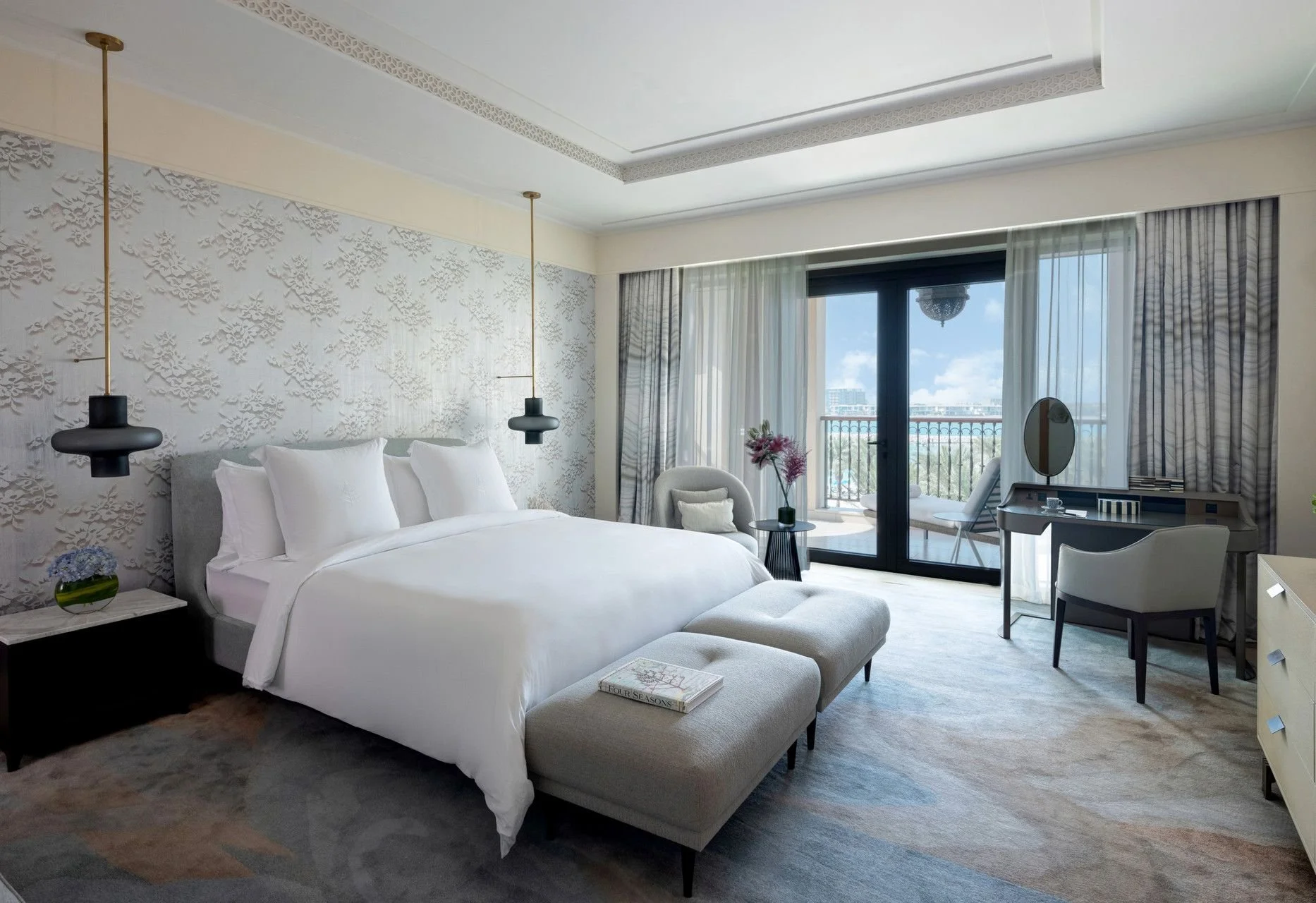 A modern hotel room with a large white bed, decorative wall panel, pendant lights, a sitting area with flowers, and a balcony with city view.