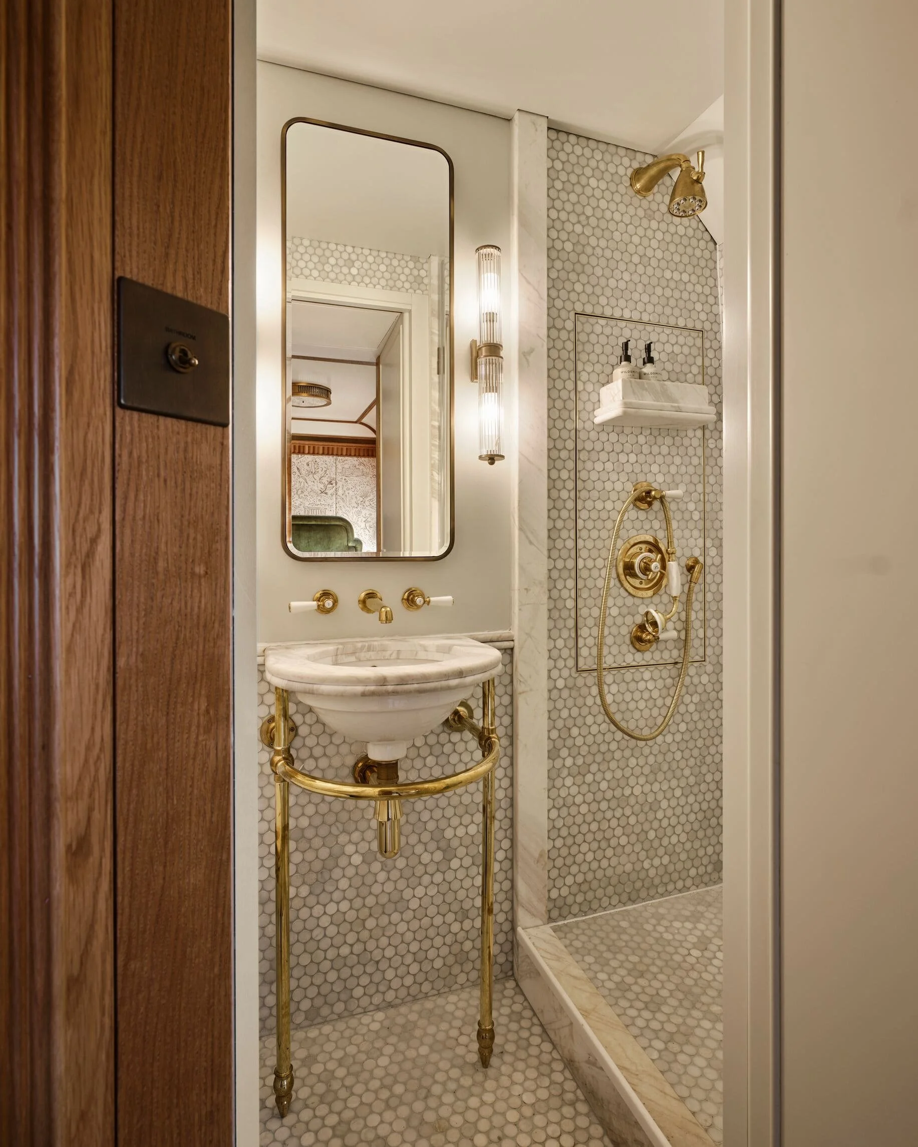 A bathroom with a vanity area containing a white marble countertop and vessel sink supported by 'T' shaped gold metal legs. The wall has small white hexagonal tiles, and the shower area features a gold showerhead and fixtures. A rectangular mirror is mounted by the wall, and there are two wall-mounted light fixtures. A wooden doorframe is visible on the left.