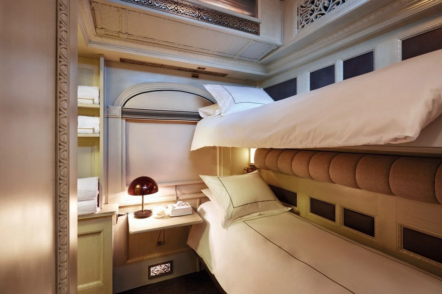 Decorative hotel bedroom with two single beds, cream-colored bedding, a small side table with a red lamp and tissue box, and a built-in shelf with folded towels. Ornate molding and paneling adorn the walls.