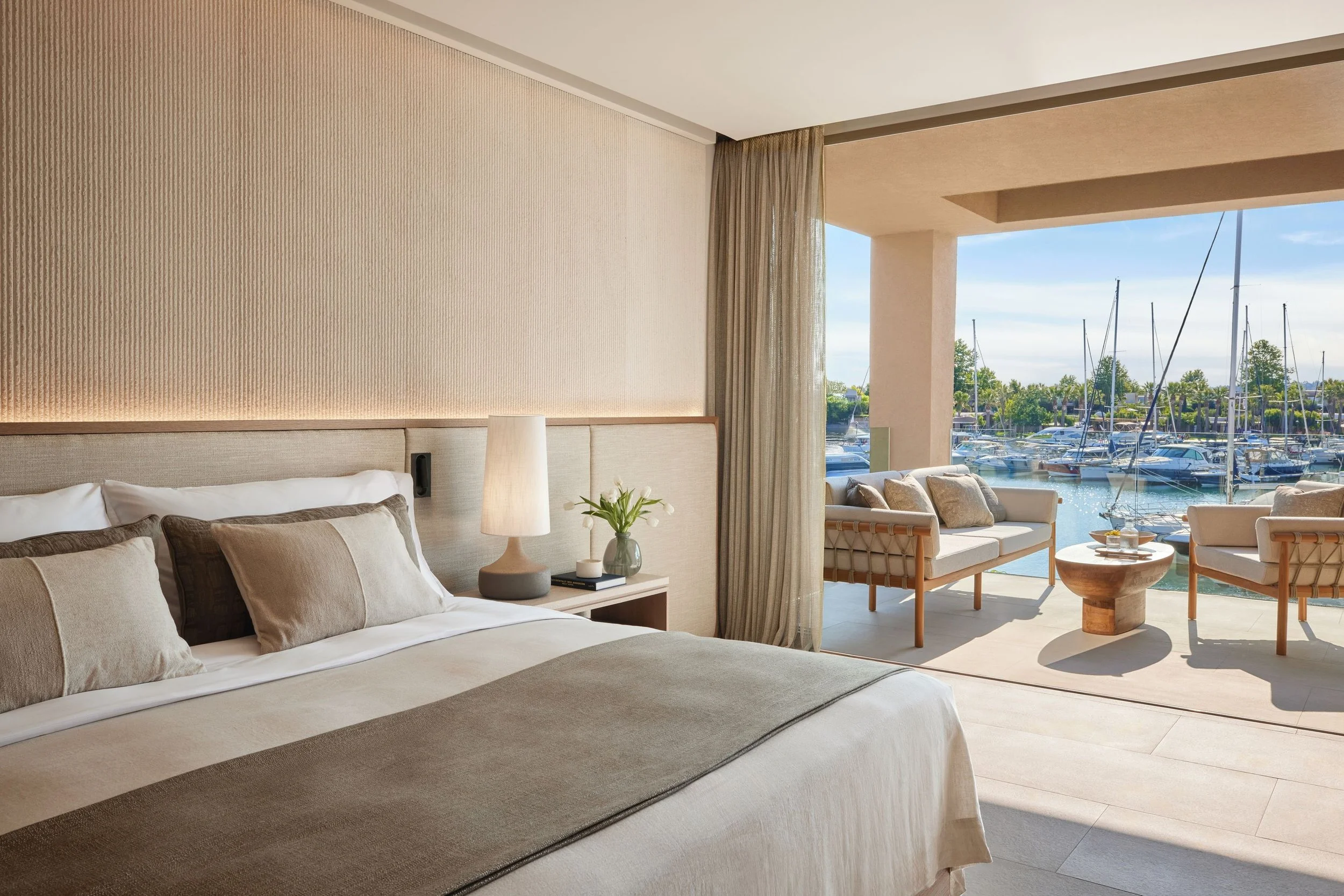 A modern bedroom with a large window opening to a marina with boats, beige curtains, a bed with beige and gray bedding, a white lamp, a plant, and seating area.
