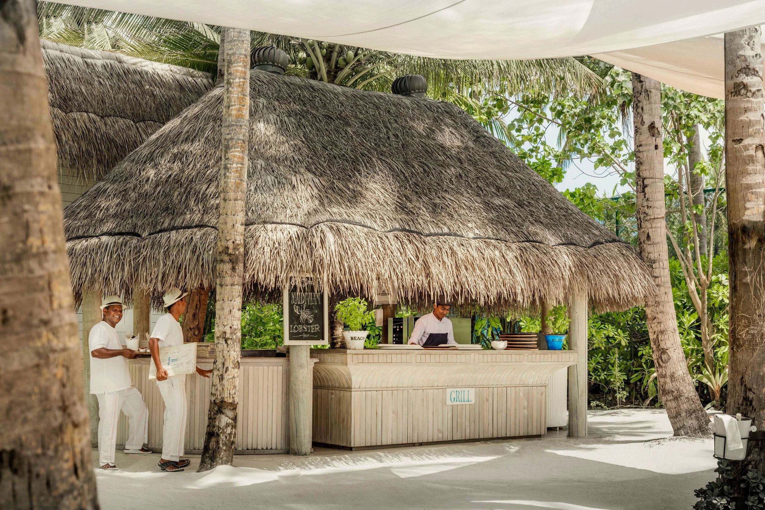 A beachside tiki hut with a thatched roof serving food and drinks. Three staff members in white uniforms are working, and there are lush green trees surrounding the hut.