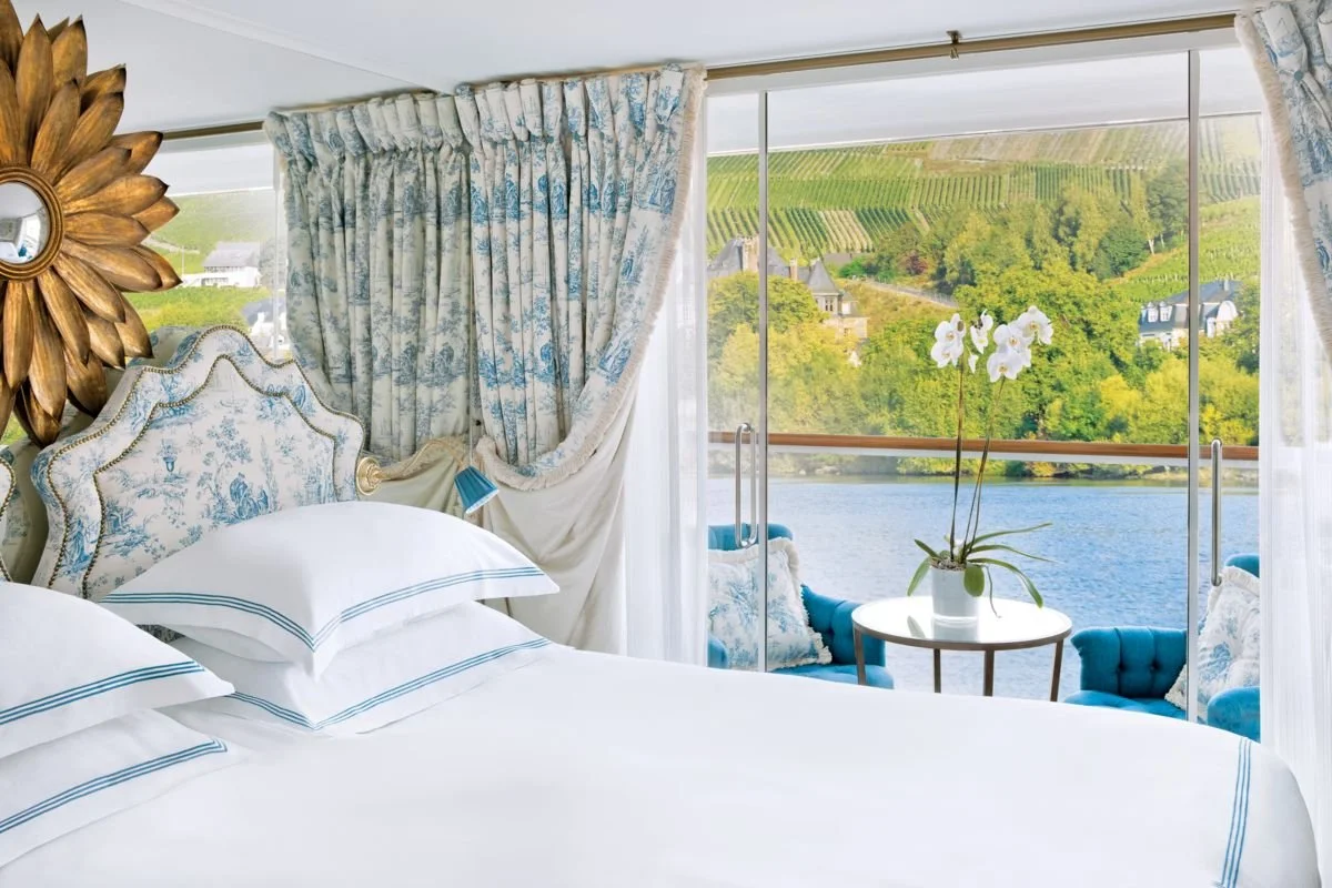 Elegant bedroom with a view of a river and lush green landscape through large glass windows, decorated with blue and white floral fabrics, a orchid plant on a small table, and blue armchairs.
