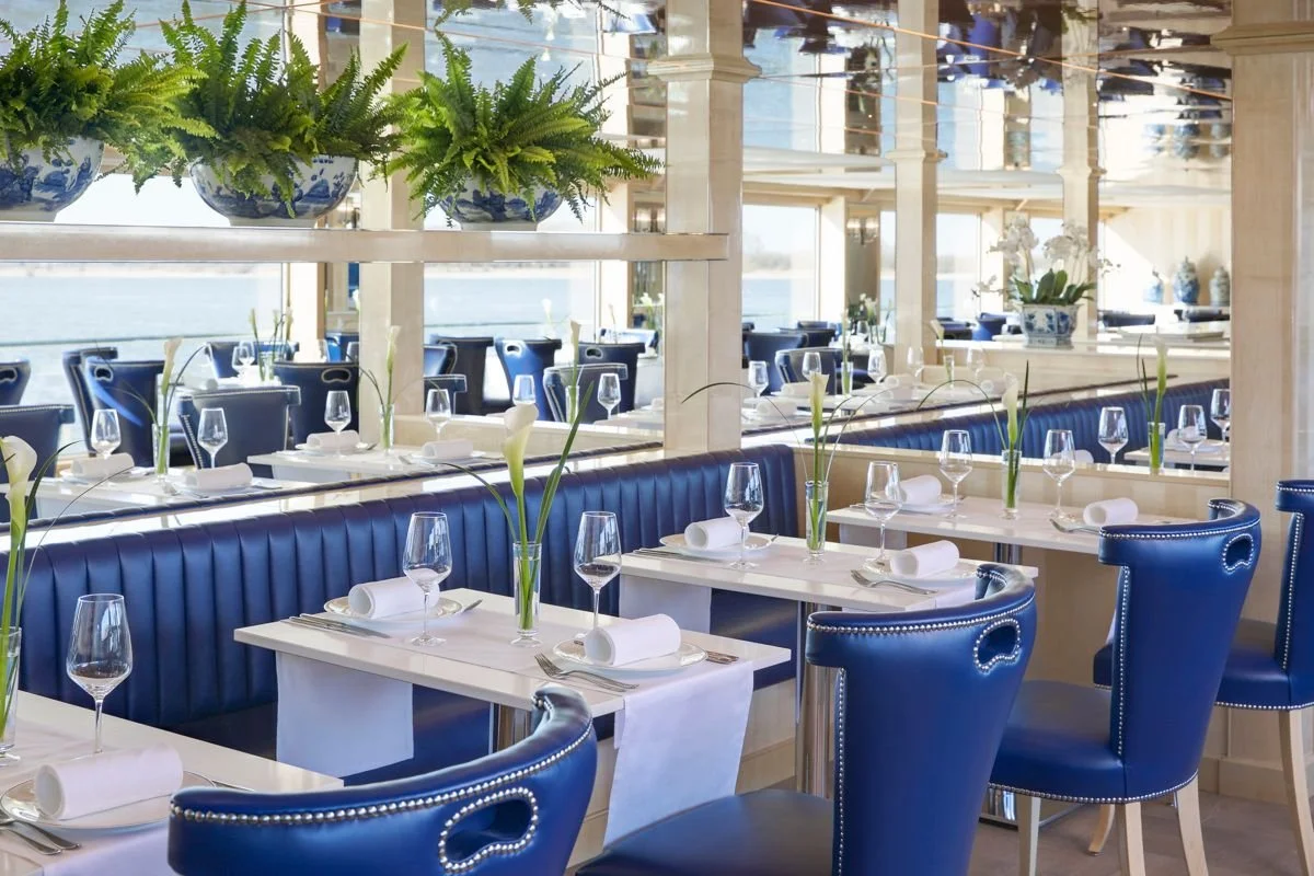 Elegant restaurant with blue chairs, white tables, wine glasses, and flower arrangements, overlooking a body of water.
