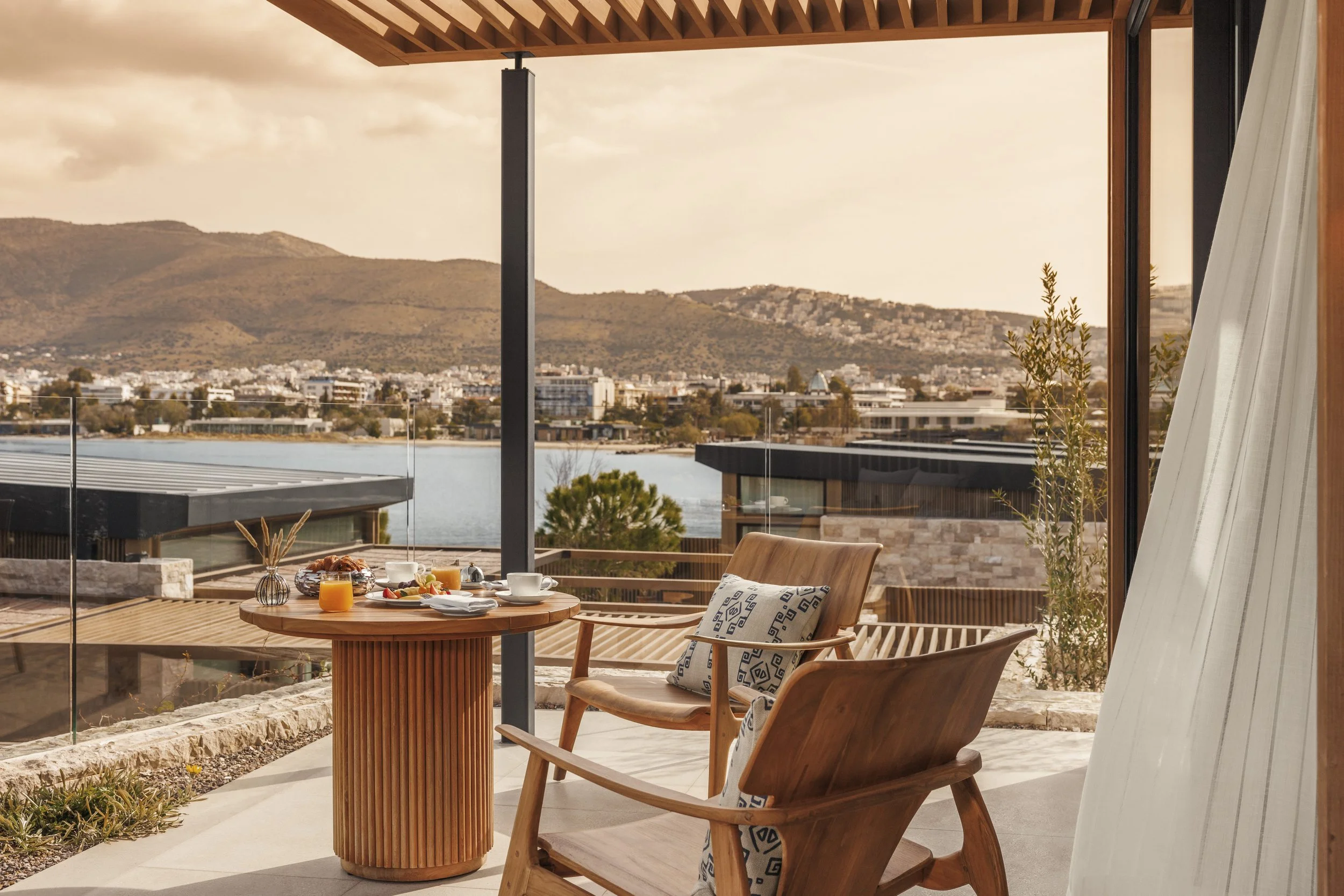 A cozy outdoor patio with a round wooden table set for breakfast, including orange juice, coffee, and food. There are two wooden chairs, one with a patterned cushion, overlooking a water body and hilly landscape through large glass doors under a wooden pergola.