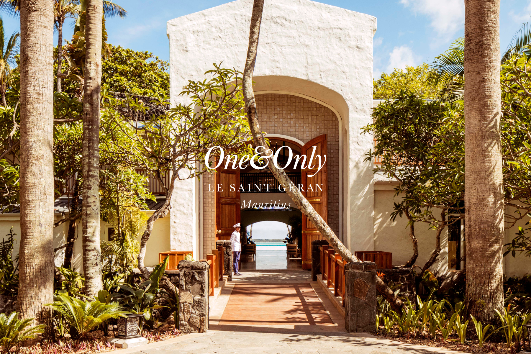 Entrance to Le Saint Gervan in Mauritius, featuring a large white textured archway, wooden door, lush tropical plants and trees, and a view of the ocean through the arch.