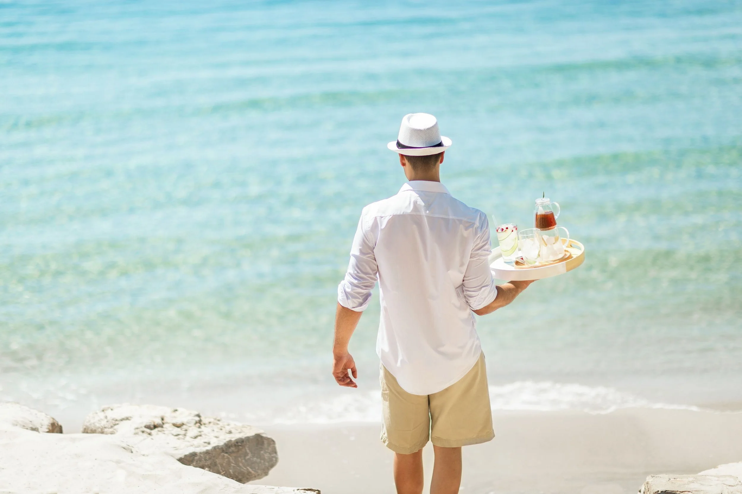 A person wearing a white shirt and beige shorts, carrying a tray with drinks, standing on a beach facing the ocean with clear blue water and rocks.