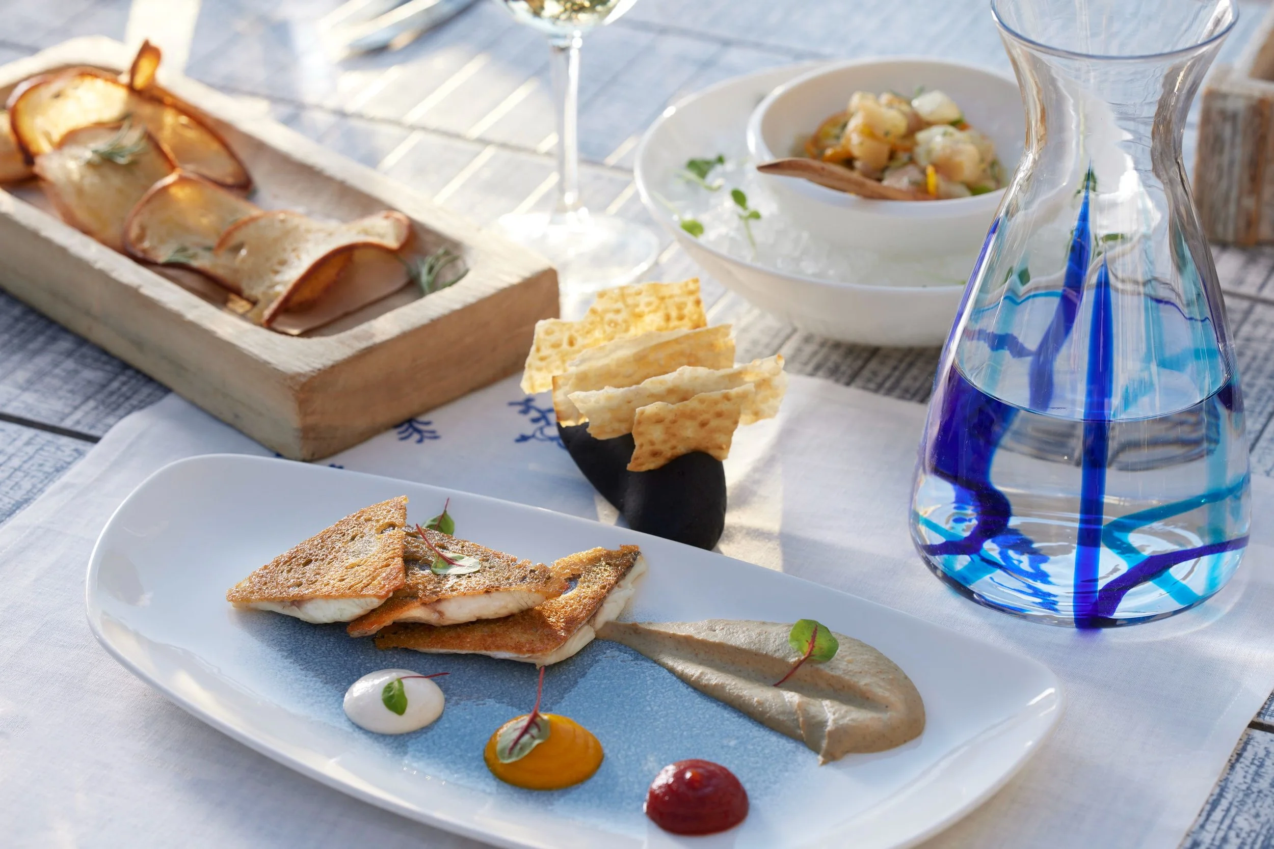 A white rectangular plate with a grilled fish fillet garnished with green herbs, served on a white cloth. There are small colorful dots of sauce, microgreens, and a streak of white sauce on the plate.