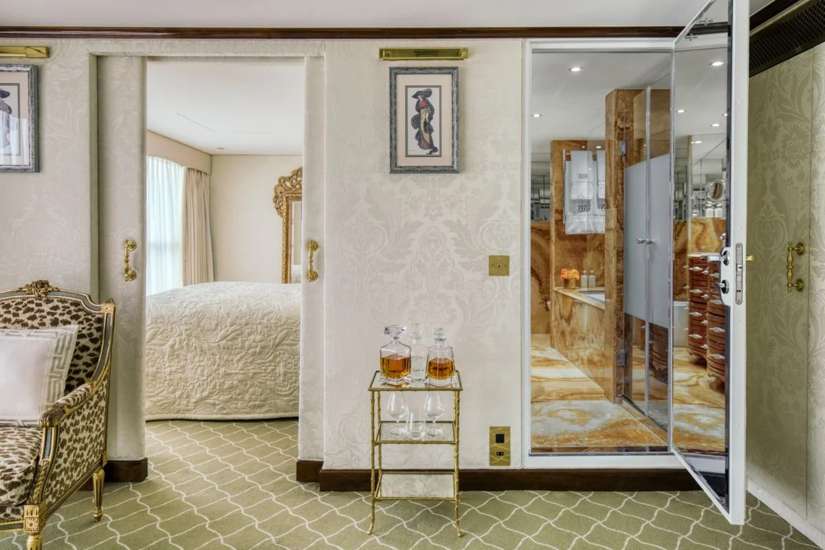 Luxurious hotel room with ornate furniture, a bed with an elaborate headboard, and a bathroom with a glass door and marble wall