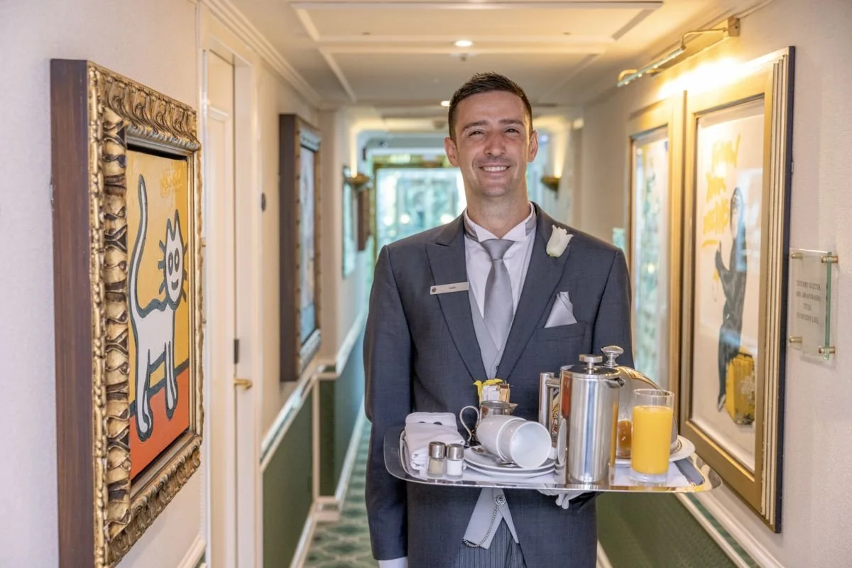 A man in a gray suit smiling and holding a tray with breakfast items in a hallway decorated with colorful art.