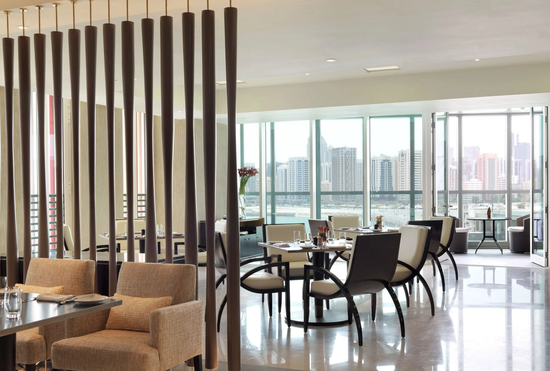 Elegant restaurant with cream-colored chairs, dark tables set with glasses and utensils, large windows showing a city skyline, and a decorative wooden partition.