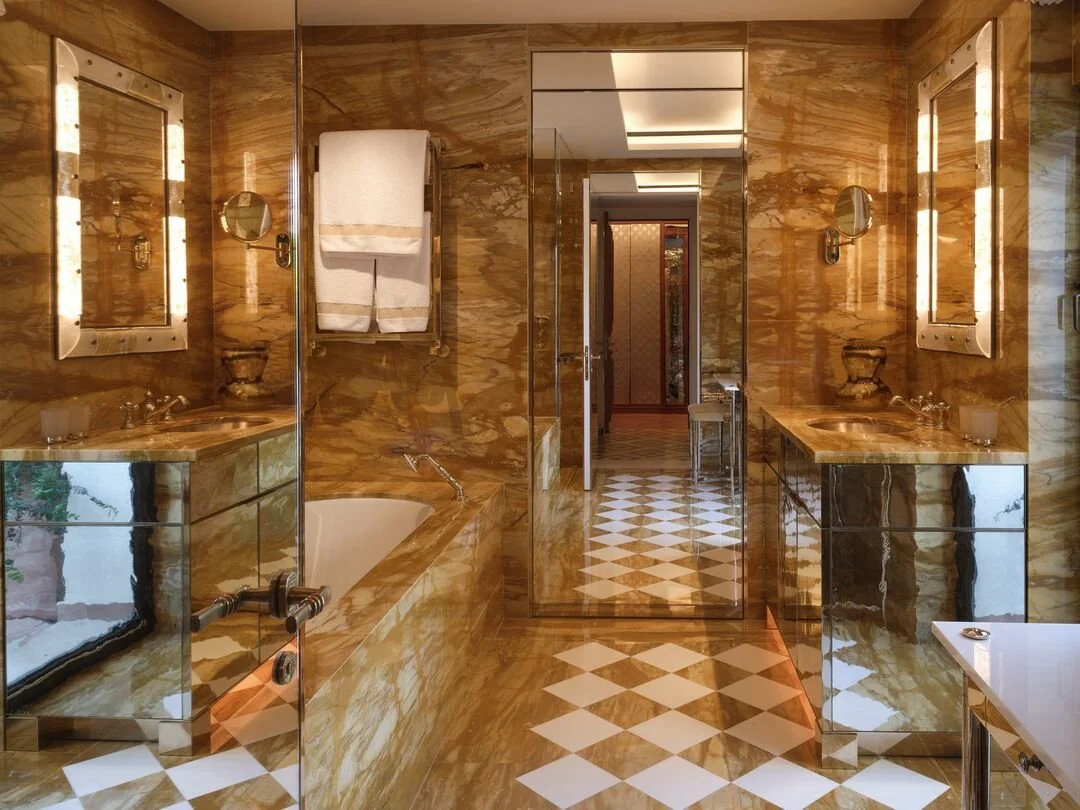 Luxurious bathroom with marble walls and flooring, double sinks with large mirrors and lighting, a corner bathtub, and a full-length mirror reflecting an adjacent hallway.