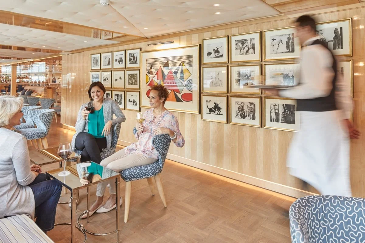 Three women sitting and talking in a stylish restaurant or lounge, with a waitress in uniform serving drinks, and framed black-and-white artwork on the wood-paneled wall behind them.
