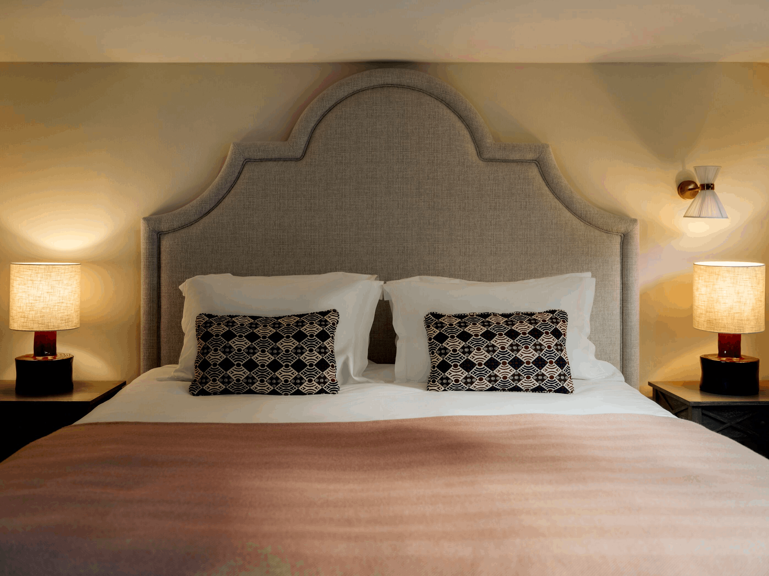 View of a neatly made bed in a hotel room with beige headboard, white pillows with black and white patterned pillowcases, and bedside tables with lamps on each side. The lighting is warm and cozy.