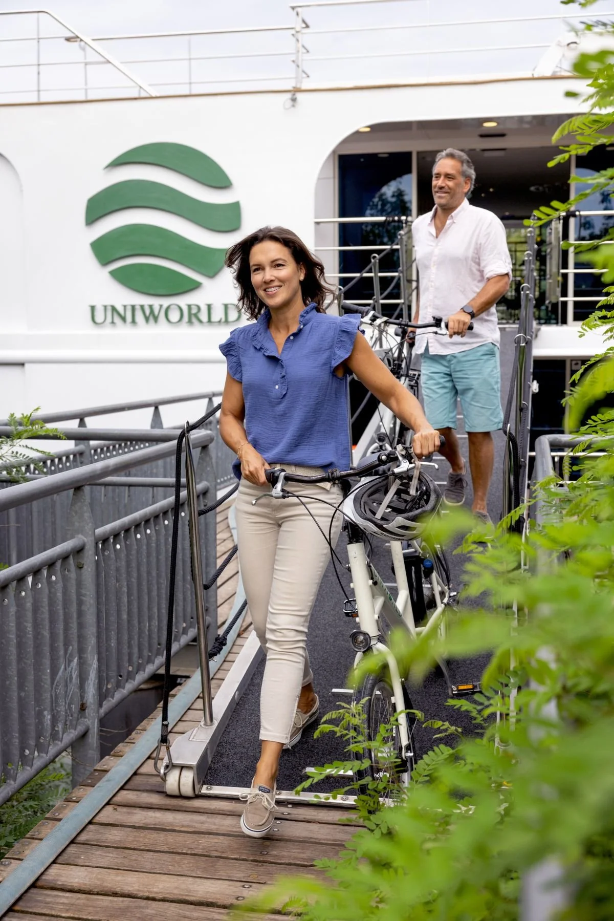 Two adults, a woman and a man, are riding bicycles down a ramp outside a building with a logo that says "UNIWORLD." The woman is in the foreground, smiling, wearing a blue blouse and beige pants, holding a bicycle. The man is in the background, smiling, wearing a white shirt and light shorts, also holding a bicycle. There are green plants and trees around them.