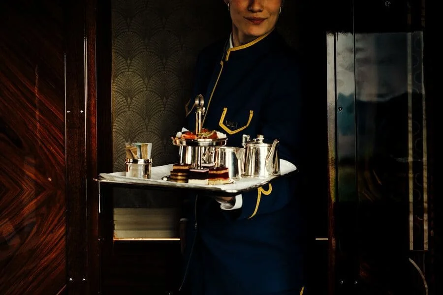 A woman in a dark uniform with gold trim holds a silver serving tray with desserts and tea set in an elevator.