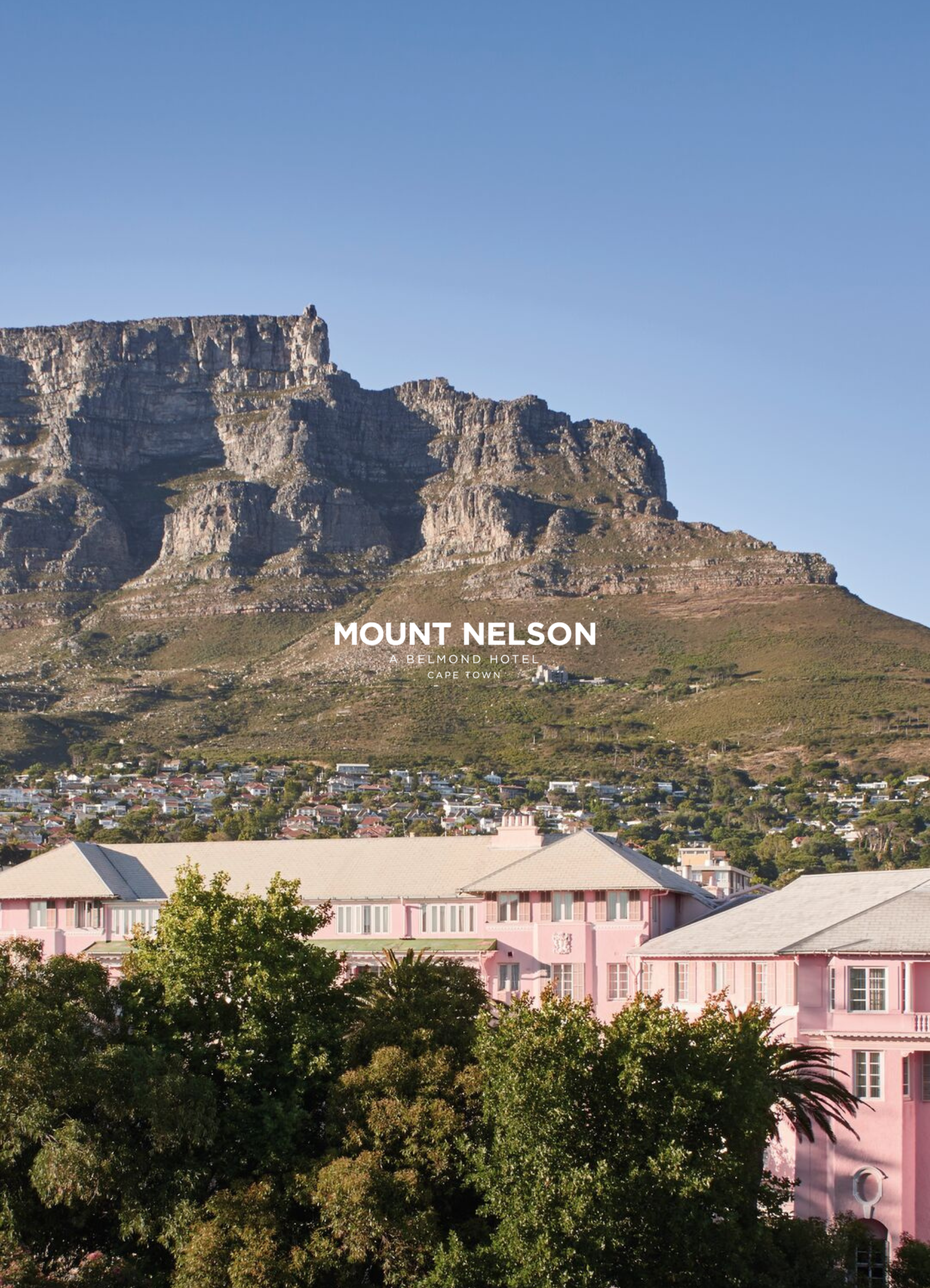 View of Table Mountain with a clear blue sky, a pink hotel building with a gray roof in the foreground, and lush green trees.