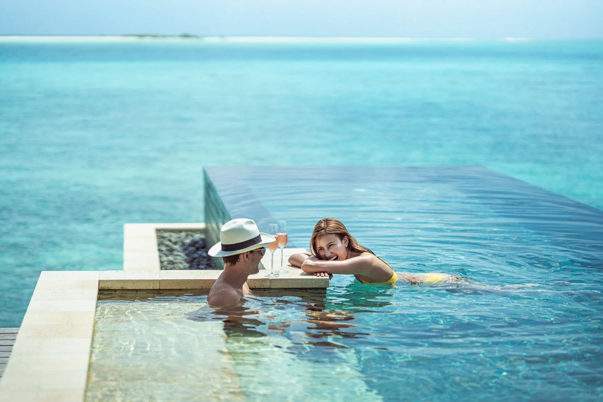 A man and woman relaxing in a swimming pool by the ocean, with two glasses of rosé wine and one with a straw on the pool's edge, the woman smiling at the man, wearing a yellow swimsuit and a wide-brimmed hat.
