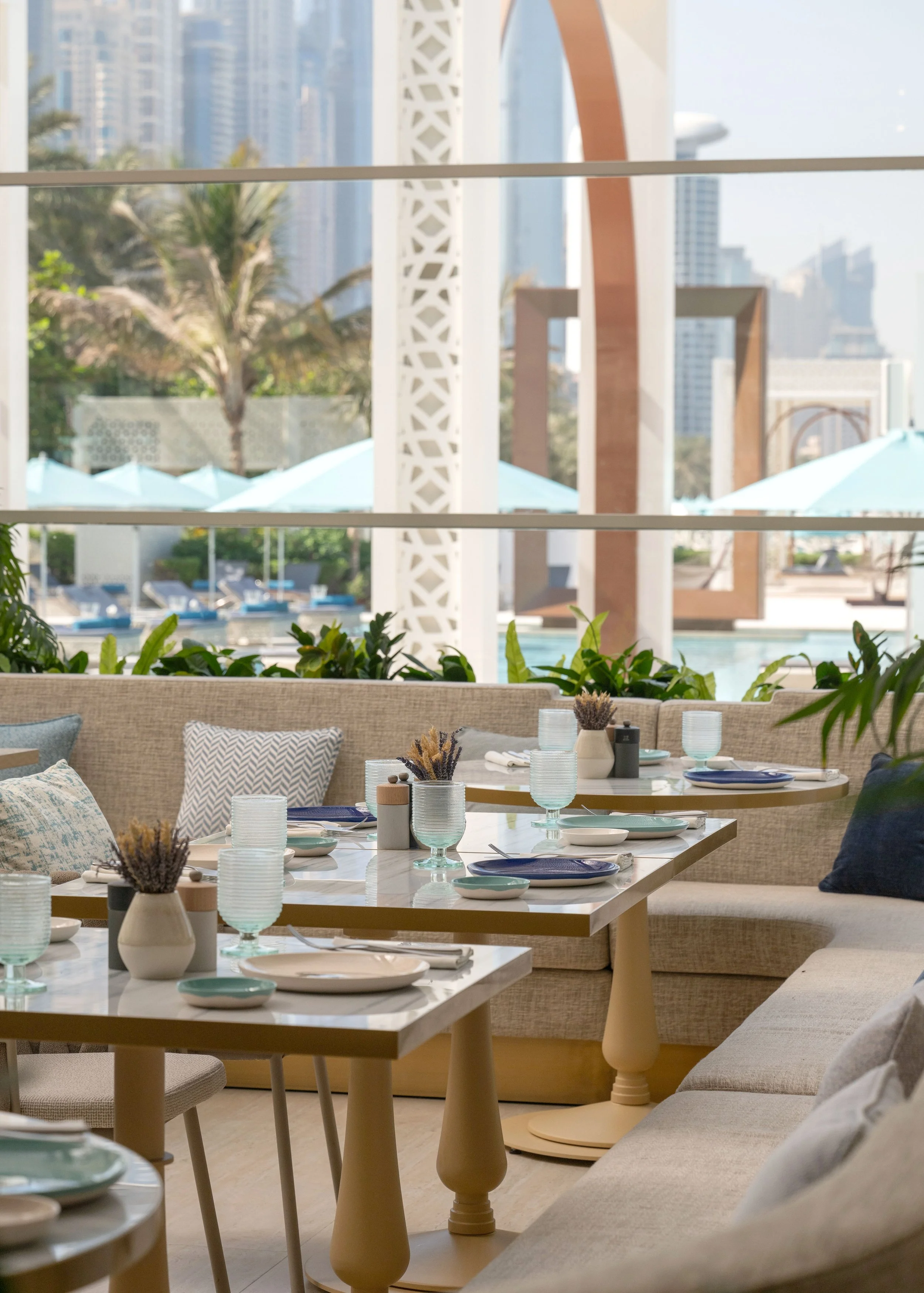 A dining area with tables set with plates, glasses, and napkins, surrounded by beige seating and cushions, with large windows showing an outdoor swimming pool, palm trees, and city high-rise buildings in the background.