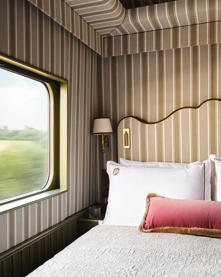 Interior of a train room with striped wallpaper, a window showing blurred scenery, a bed with white pillows and a pink bolster pillow, and a wall-mounted light fixture.