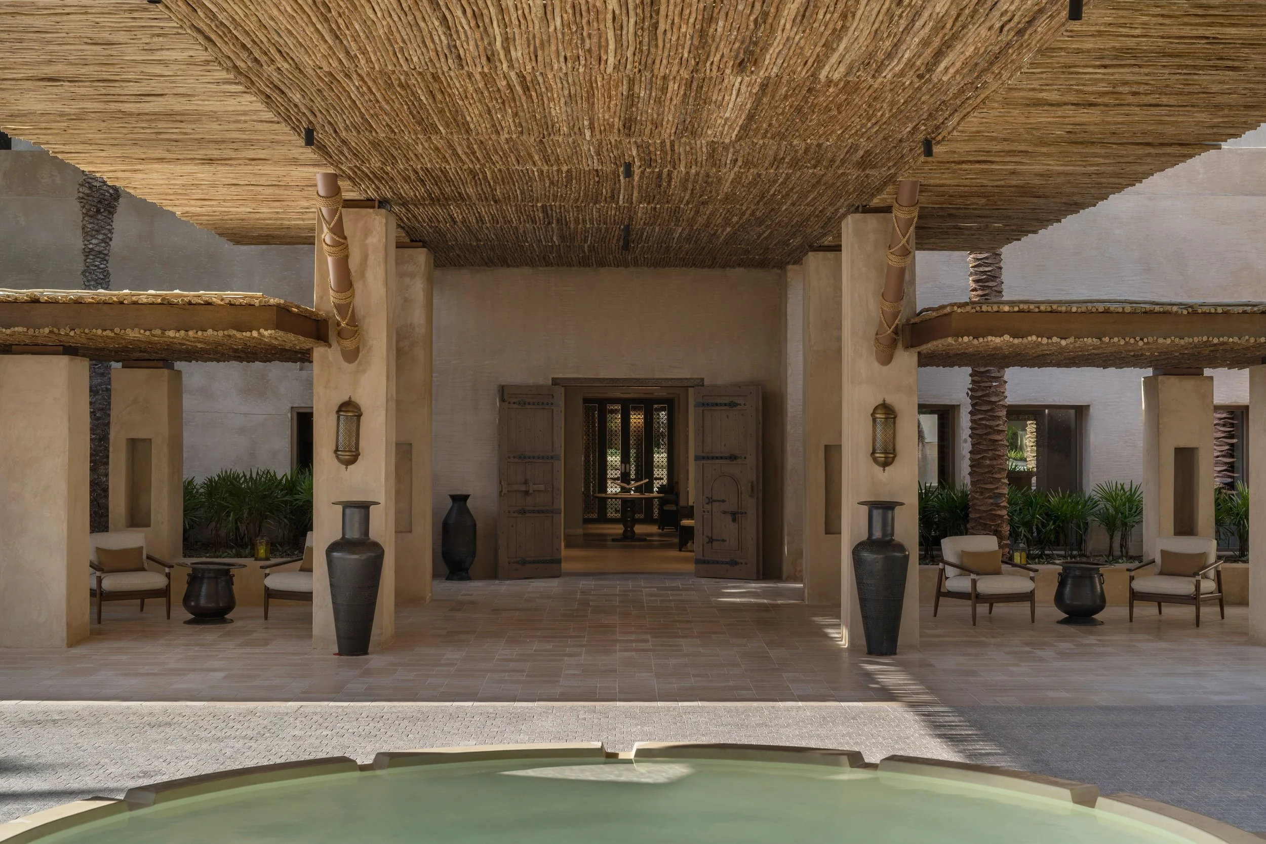 Front view of an outdoor lounge area with beige chairs, black planters, and a thatched roof overhead, leading into a building with traditional wooden doors.