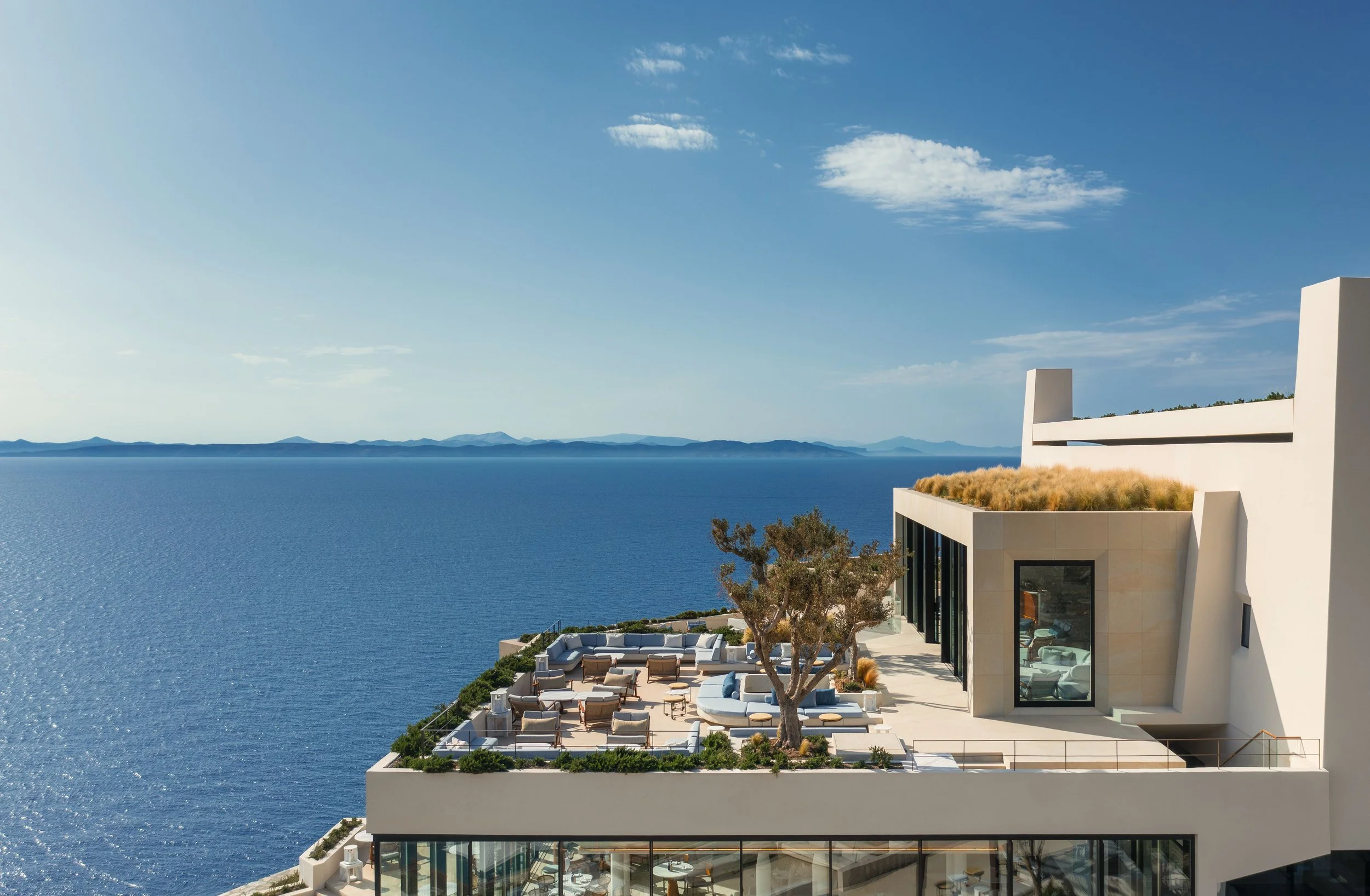 Modern white building with rooftop terrace overlooking blue ocean and distant islands under a partly cloudy sky.