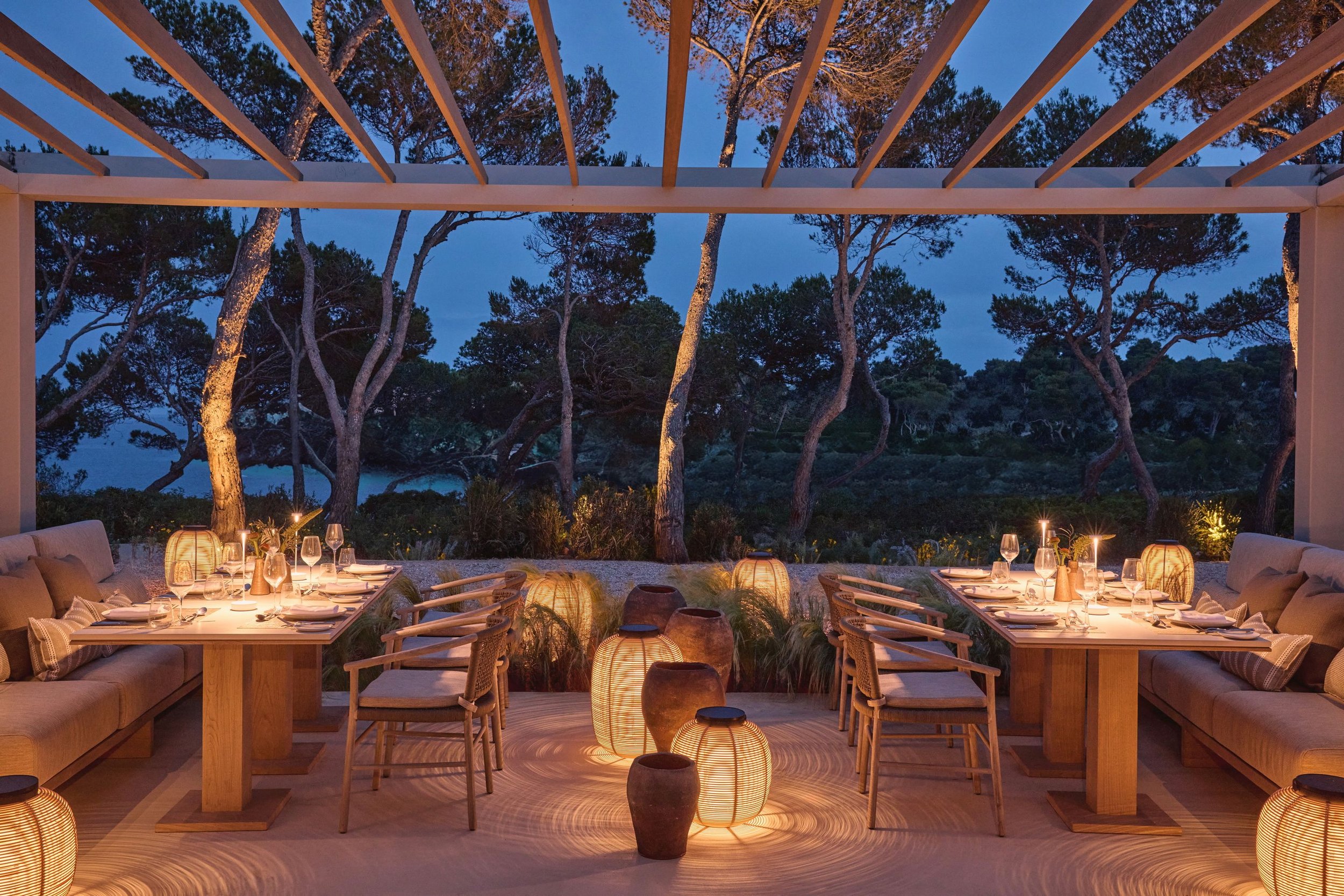 Outdoor dining setup at dusk with tables and chairs surrounded by lanterns and trees in the background.