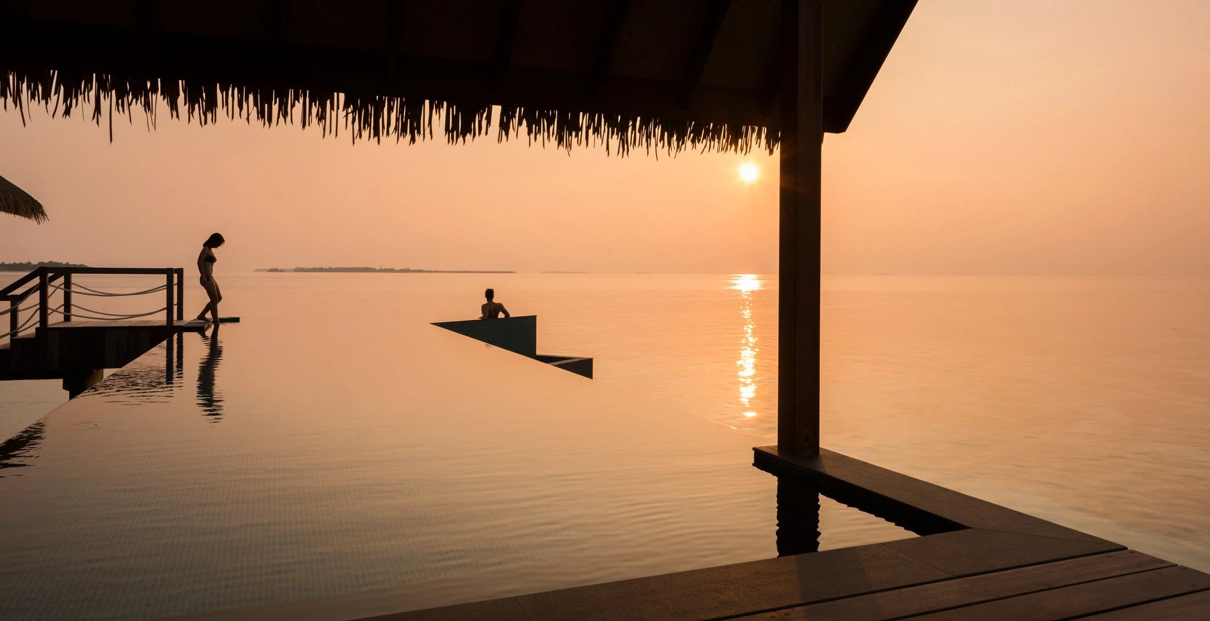 Silhouette of two people by a sunset over the water, with a thatched-roof structure and dock in the foreground.