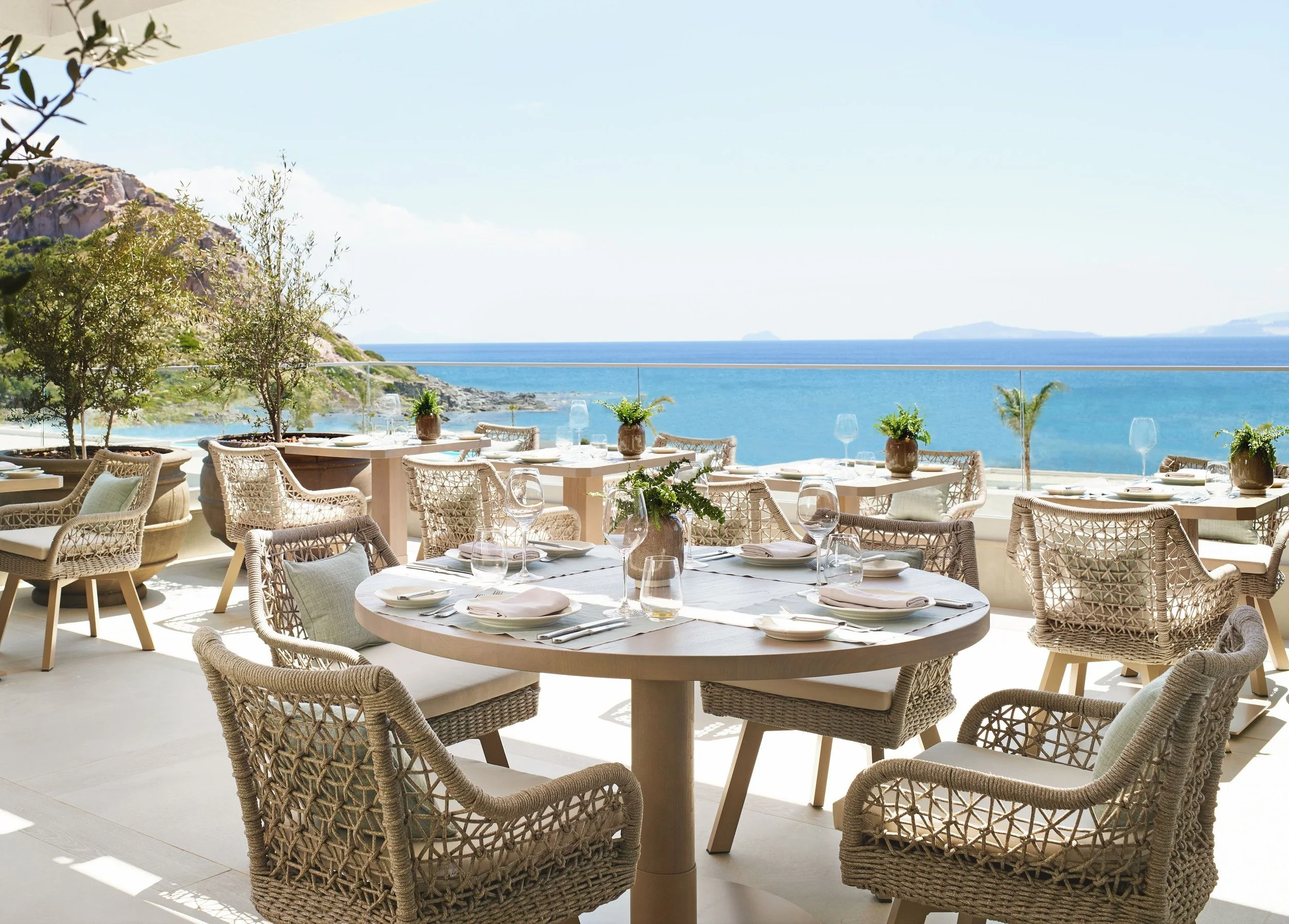 A seaside restaurant with round tables set for dining, adorned with white napkins, glassware, and potted plants, overlooking the ocean with a rocky hillside and distant islands in the background, under a clear sky.