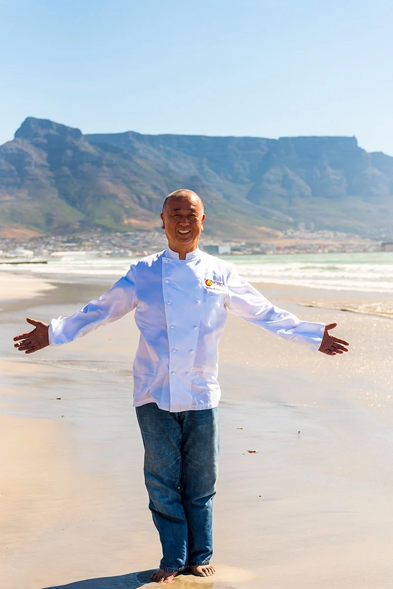 A man in a white chef's coat standing on a sandy beach with arms outstretched, with Table Mountain visible in the background.