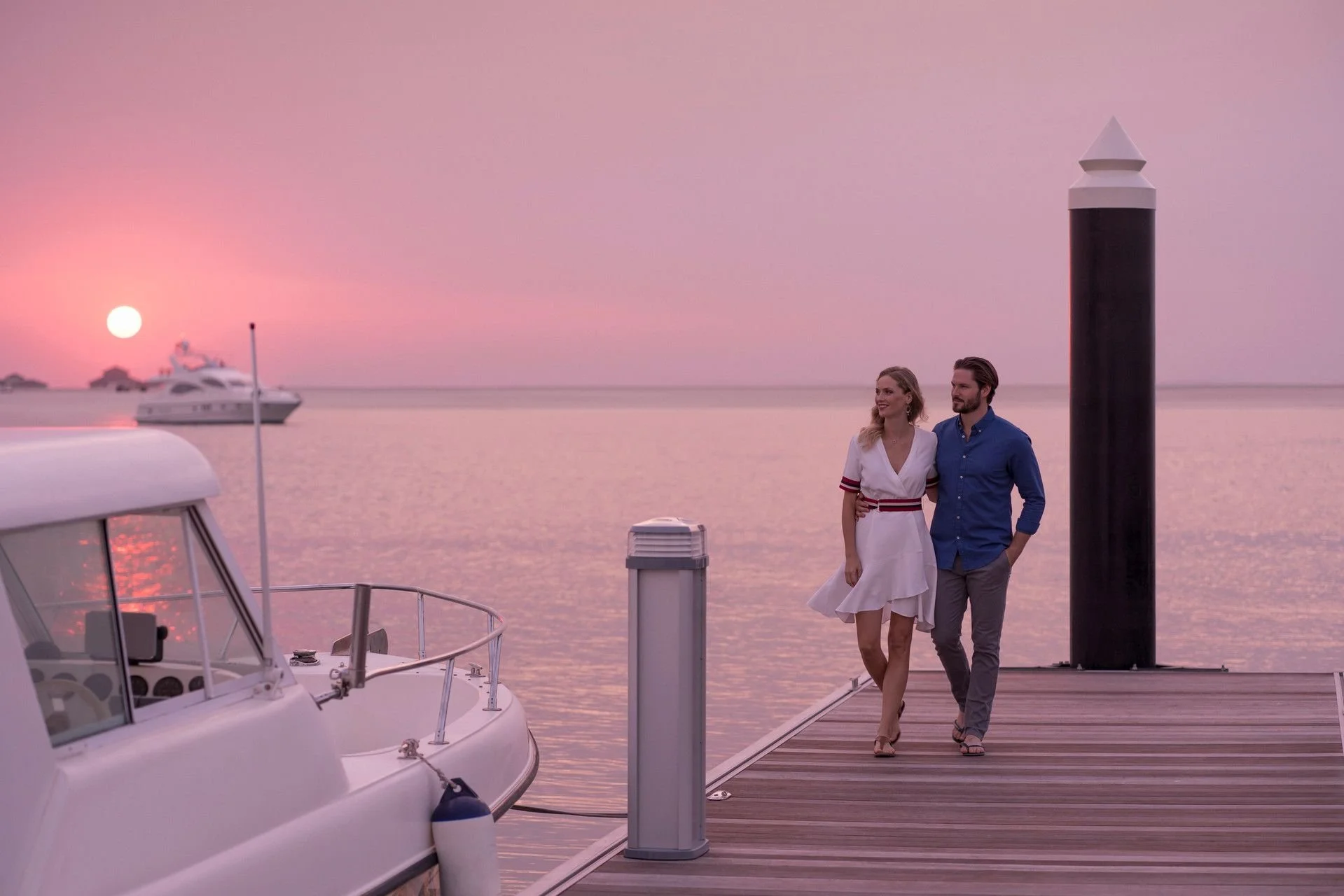 A couple walking on a dock by the water during sunset, with yachts anchored in the background and a pink sky overhead.