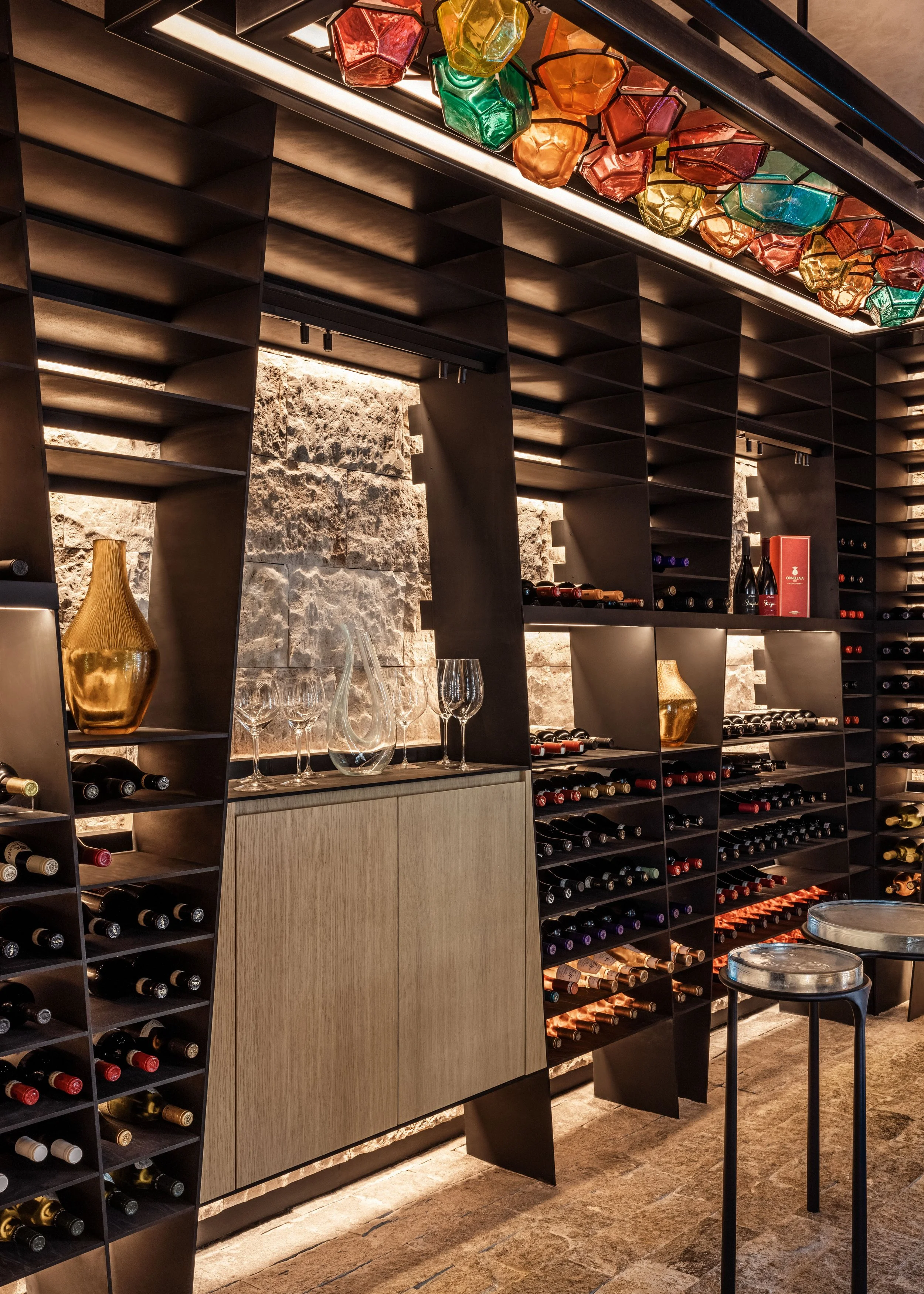 A modern wine cellar with dark wooden wall-mounted shelves filled with wine bottles. Colorful glass lanterns or decorative light fixtures hang from the ceiling. There are glassware and decorative items, including a gold vase, on a counter below the shelves. The room has warm lighting and a rustic stone wall backdrop.