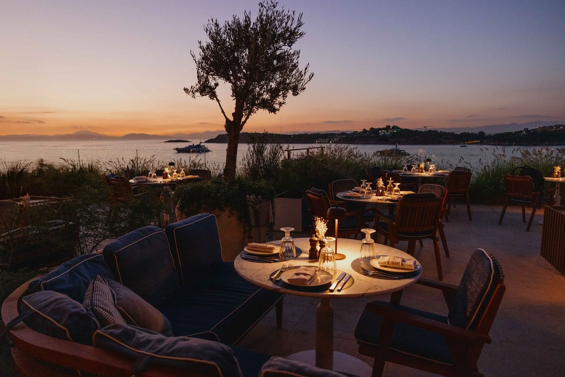 Outdoor restaurant at sunset overlooking a calm body of water with distant islands. Tables are set with glasses, plates, and cutlery, and some tables have candles. There are chairs and cushions around, with a tree in the center.