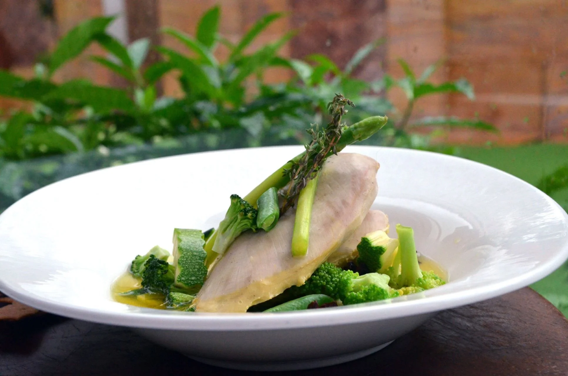 A dish with a piece of white meat, possibly chicken, topped with asparagus, herbs, and broccoli, served in a white bowl on a dark surface, with greenery and a brick wall in the background.