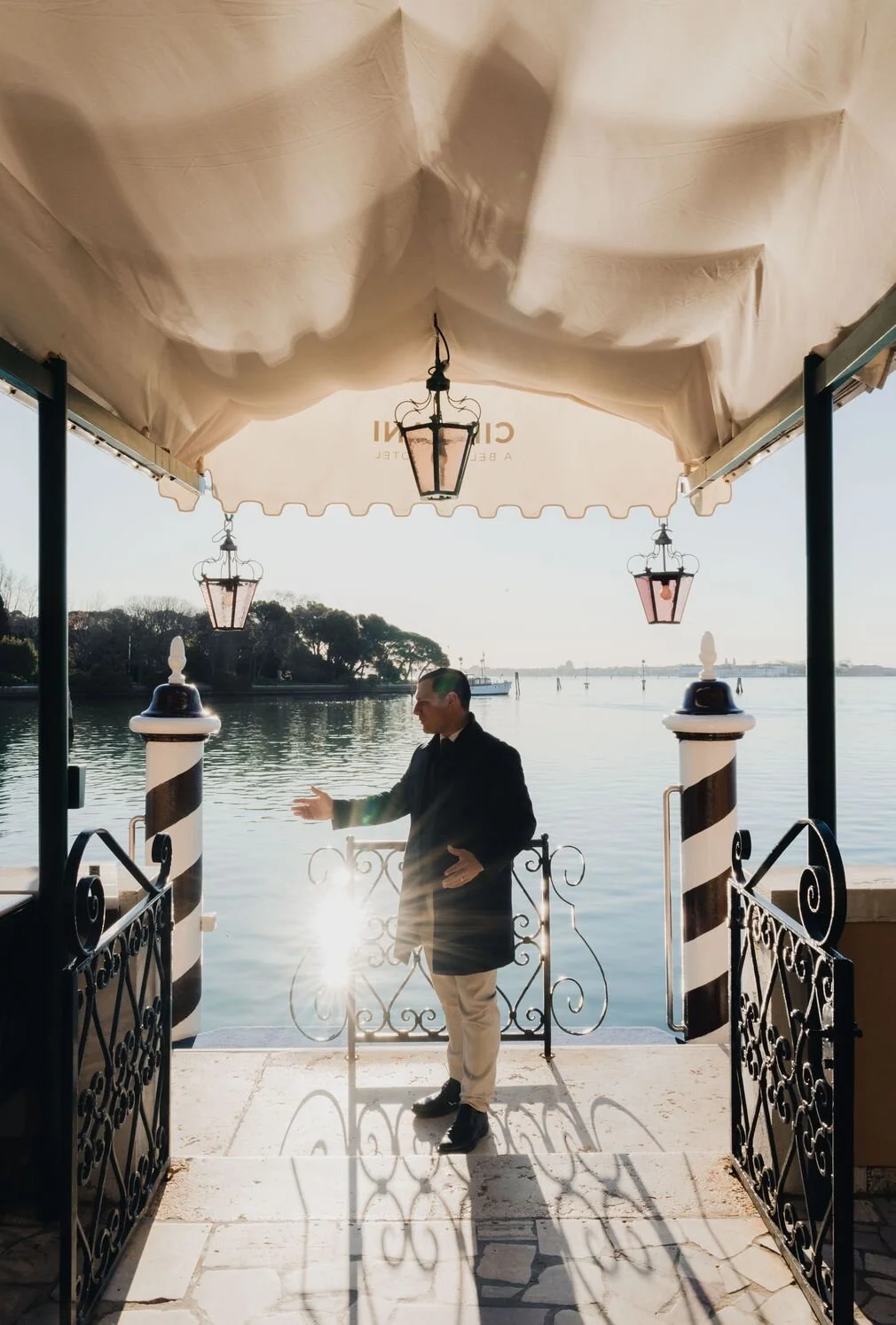 A man in a black coat and beige pants standing on a dock, smiling and gesturing with his hands, with water and boats in the background, under a white canopy with hanging lanterns.