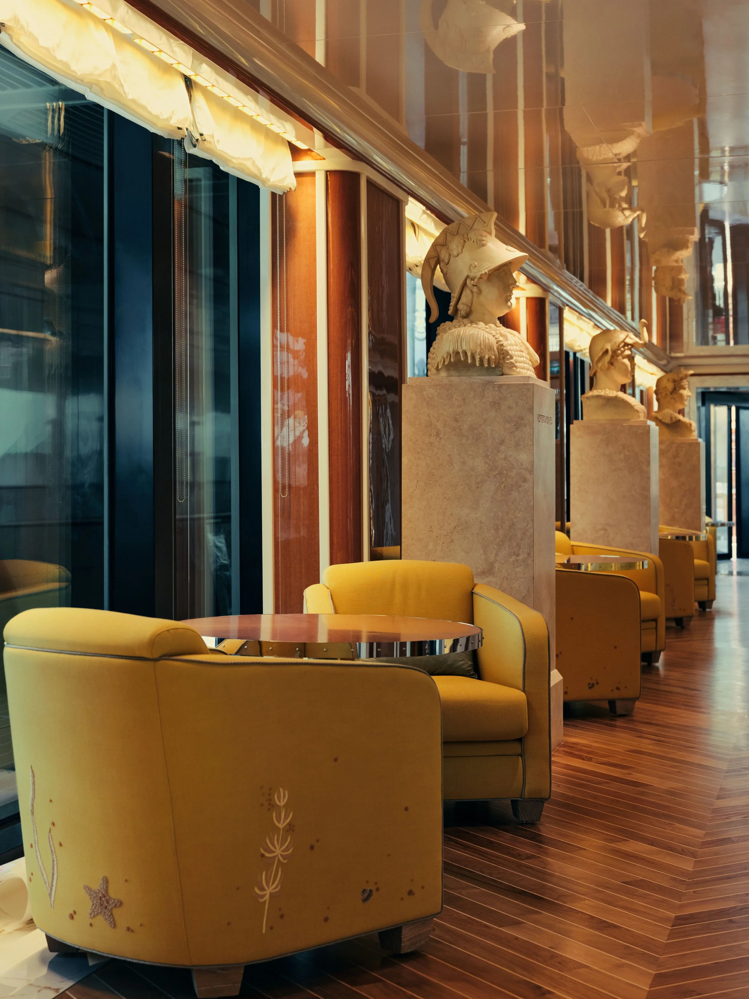 Interior of a hotel lobby or lounge area with yellow curved couches, wooden flooring, decorative bust sculptures, and mirrored walls.