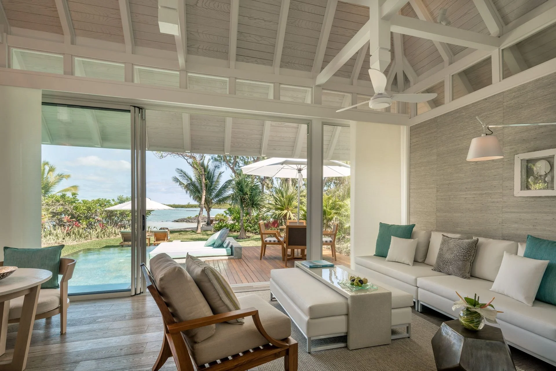 Interior view of a bright living room with large sliding glass doors opening to a tropical outdoor patio with trees, umbrellas, and water in the background. The room has a white sofa with pillows, a wooden side table, and additional chairs, with high vaulted ceilings and a ceiling fan.