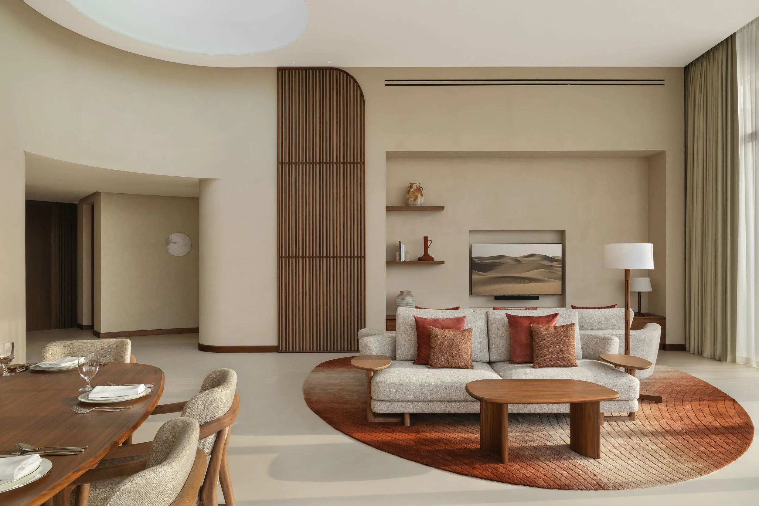 Modern living room with beige walls, wooden accents, a beige sofa with rust-colored pillows, a round coffee table, and a TV mounted on the wall. There is a curved wooden room divider, a floor lamp, and large windows with floor-length curtains.