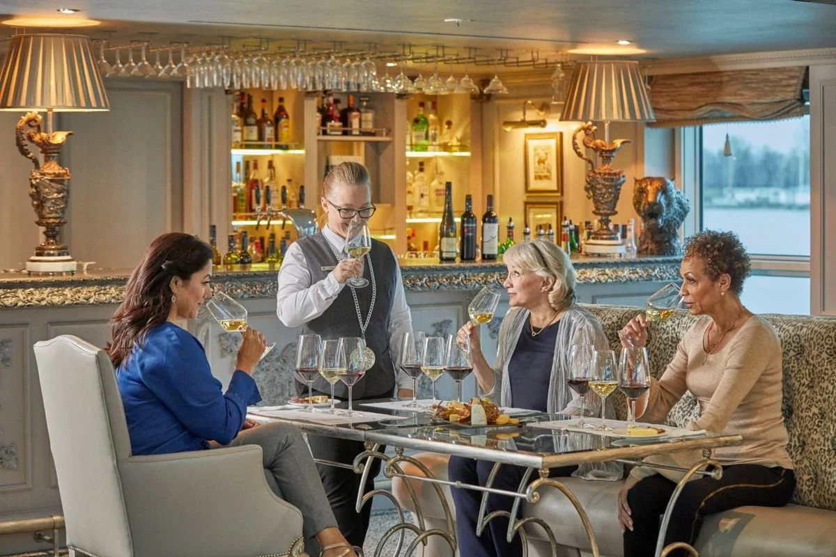 Four women sitting at a dining table in a restaurant, with a bartender serving them wine. The setting is elegant with a bar in the background, wine glasses hanging from the ceiling, and decorative items on the bar counter.