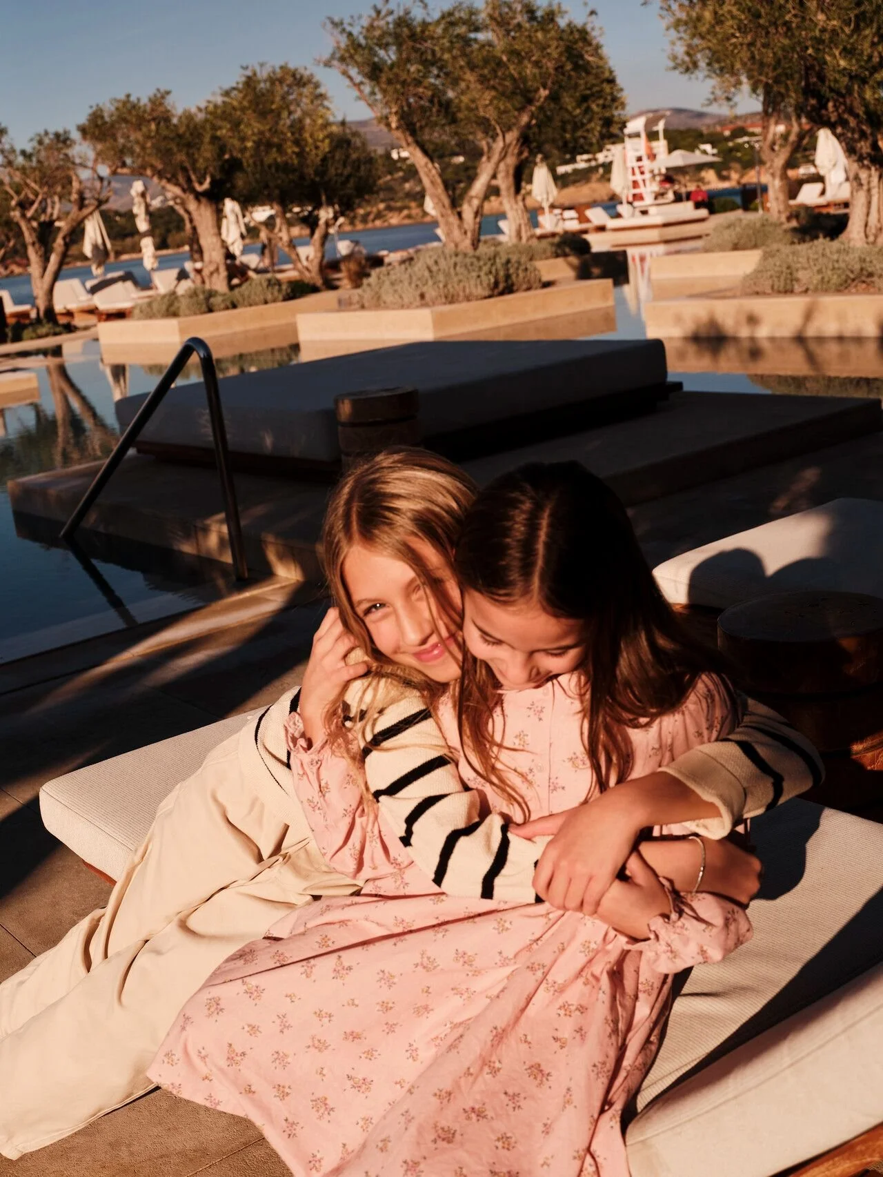 Two young girls sitting closely together on a lounge chair outdoors, smiling and sharing a hug during sunset with a pool and trees in the background.
